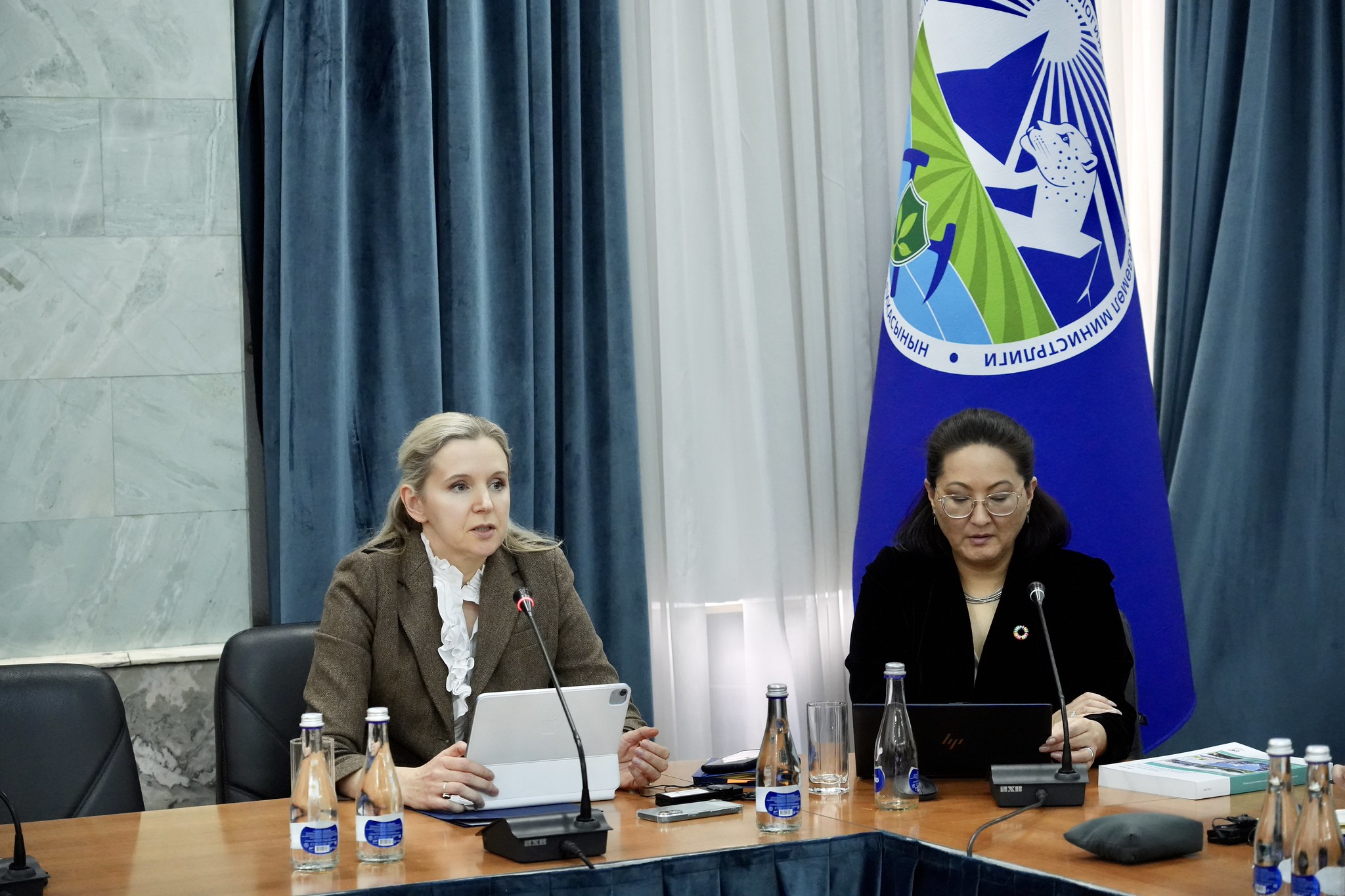Two women sit at a formal conference table with papers and water bottles; blue curtains and a flag behind.