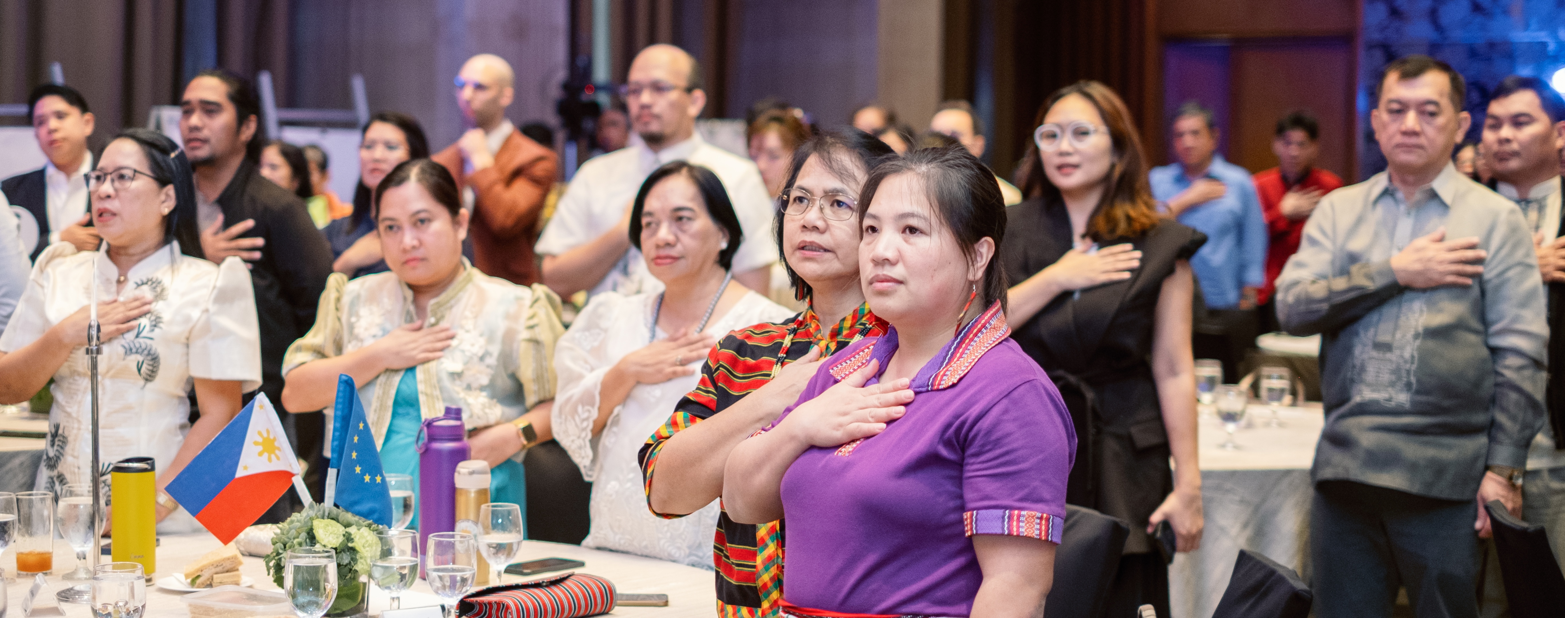 Audience at a formal gathering; foreground woman in purple claps amid colorful flags.