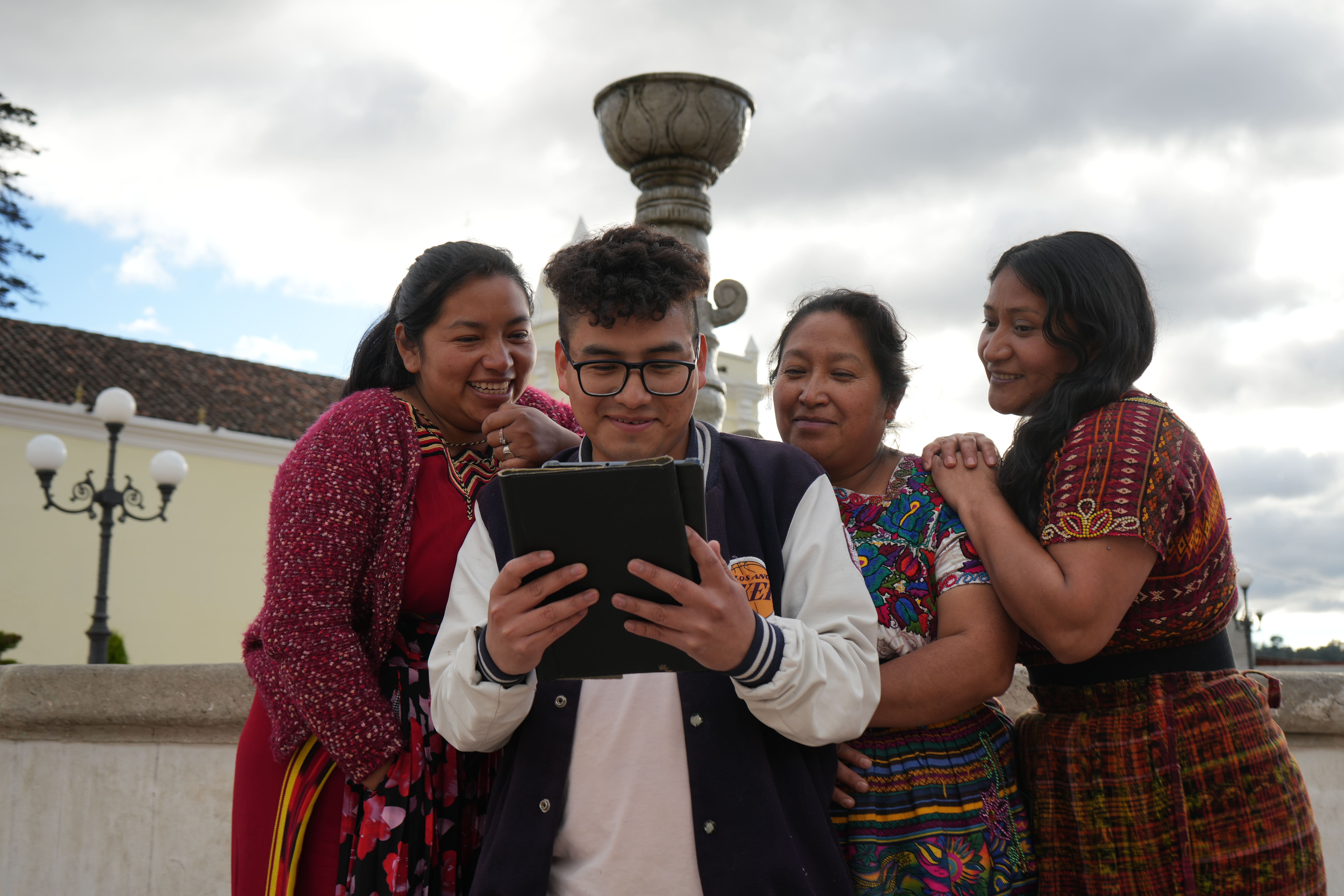 Four friends in colorful traditional clothing smile at a tablet outdoors beside a stone fountain.