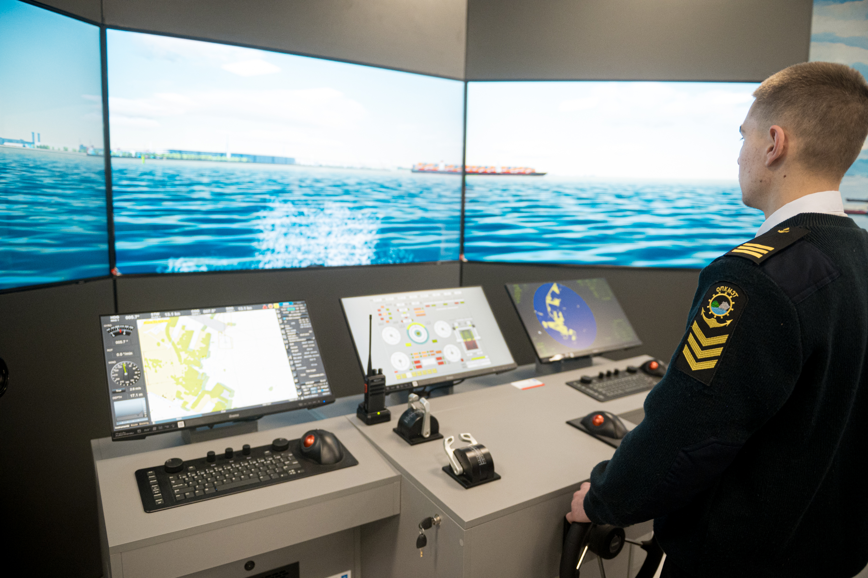 Naval officer in uniform at a ship's bridge with multiple navigation screens and a sea view.
