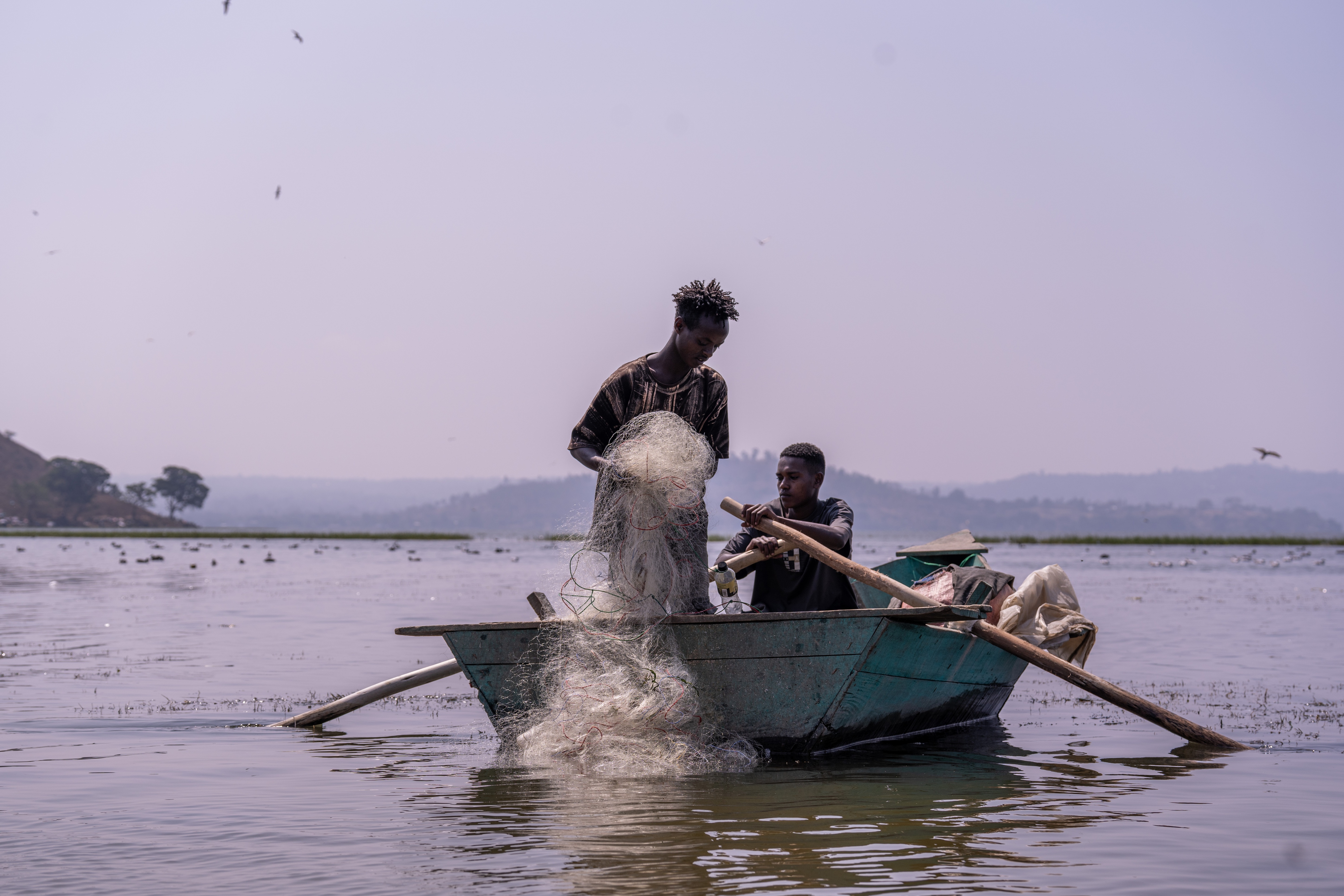 Photograph: man standing at the bow of a blue boat on calm water, splashing water as another person sits