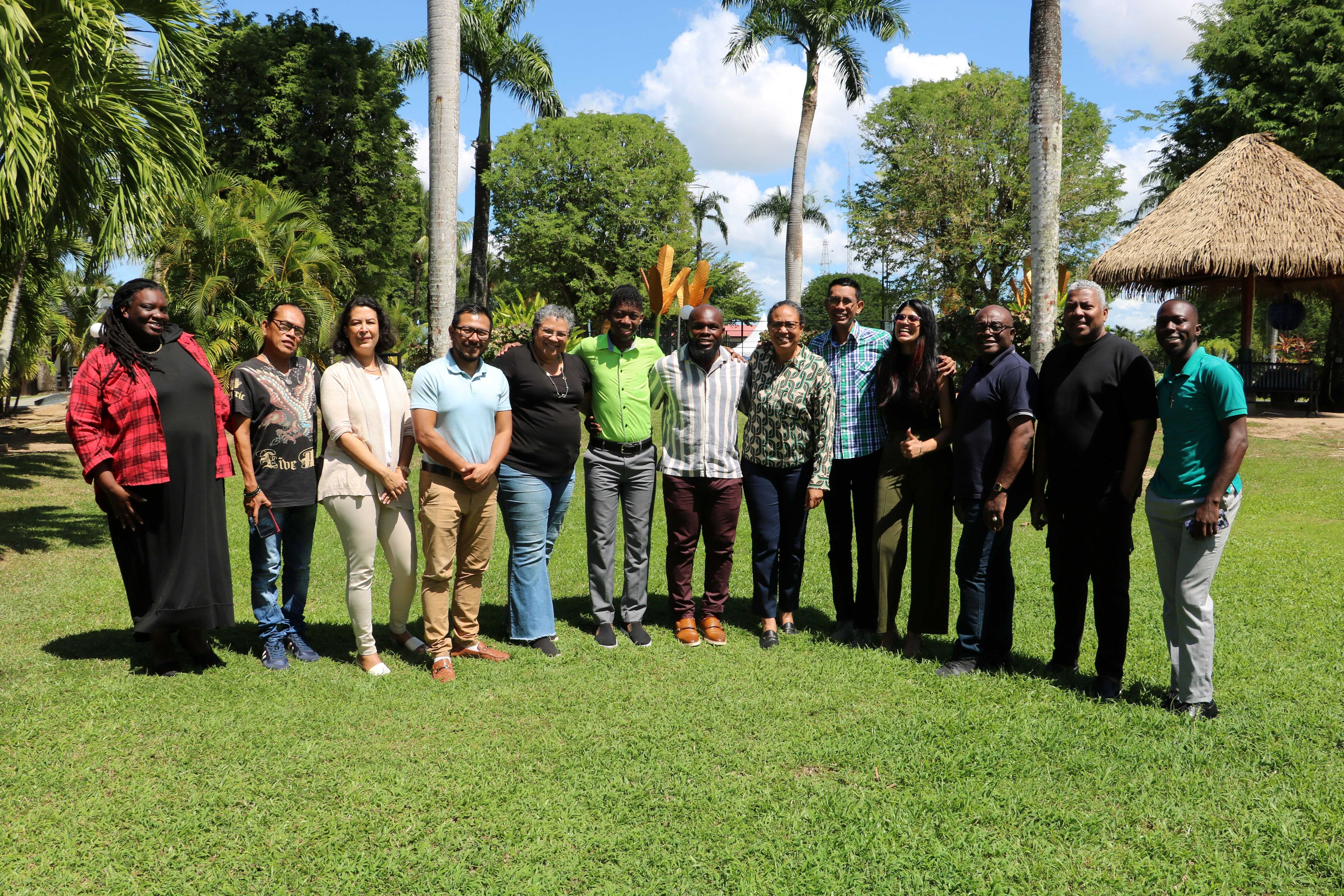Group of people standing on a grassy lawn with palm trees and a thatched hut.
