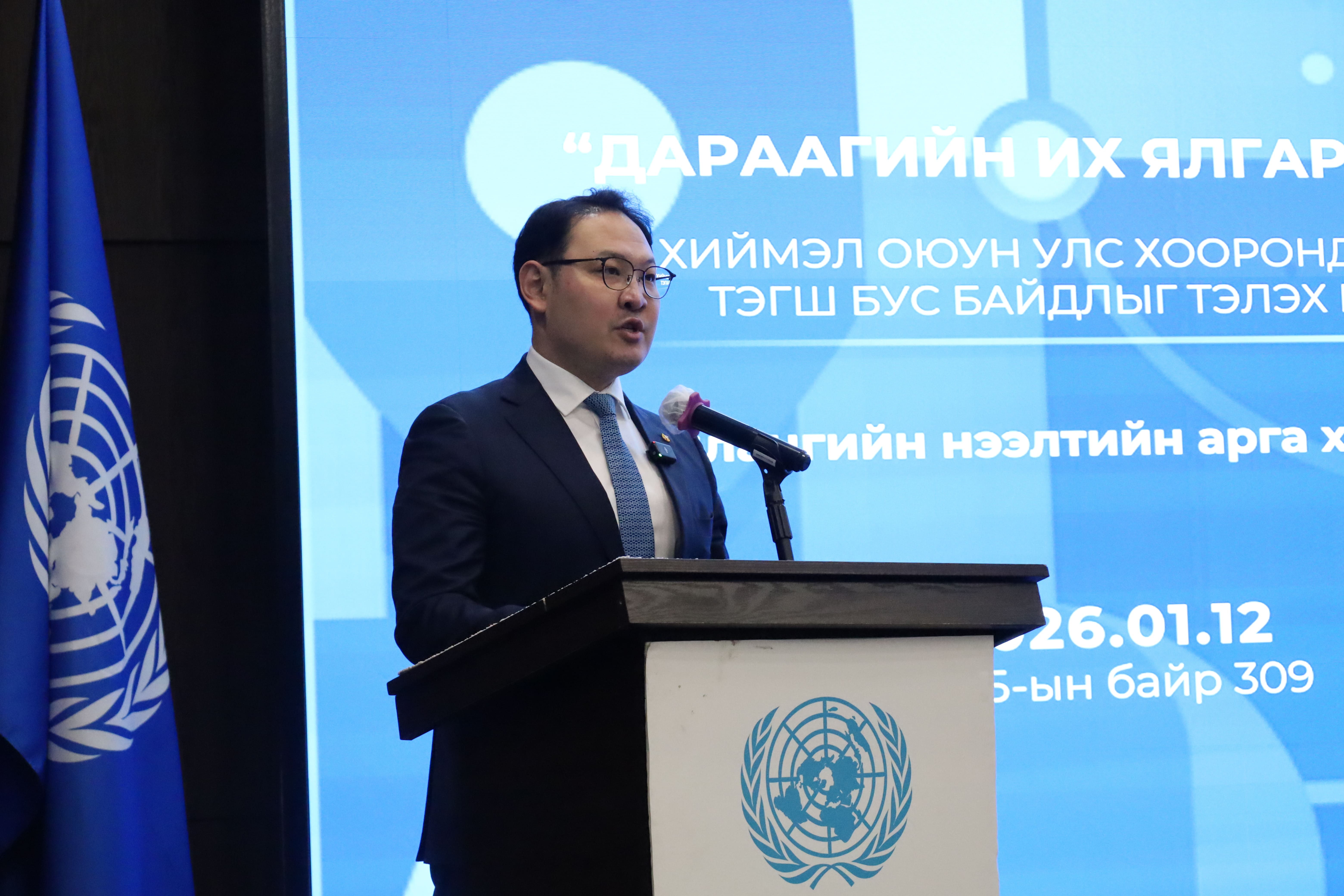 Man in suit speaks at a podium with UN emblem, blue backdrop, and UN flag.