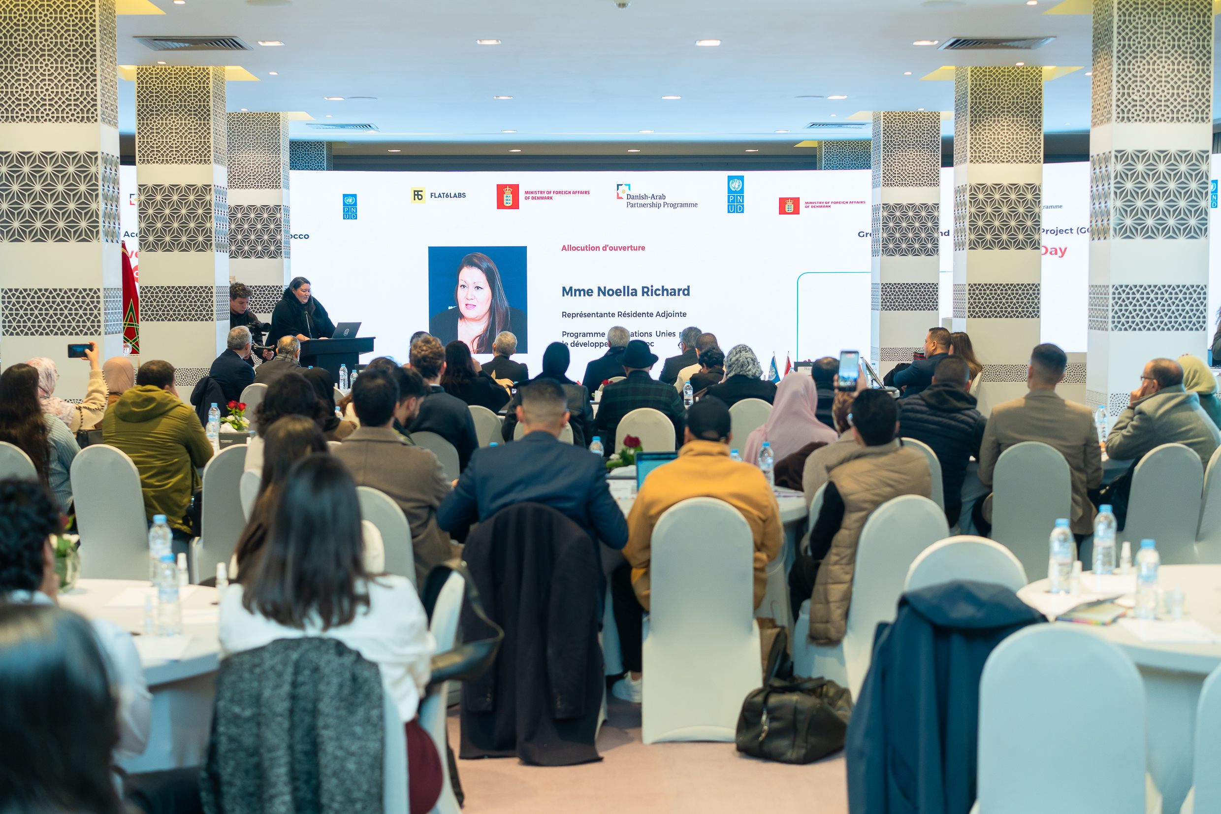 Conference hall with audience seated at tables watching a presenter on stage with a large screen.