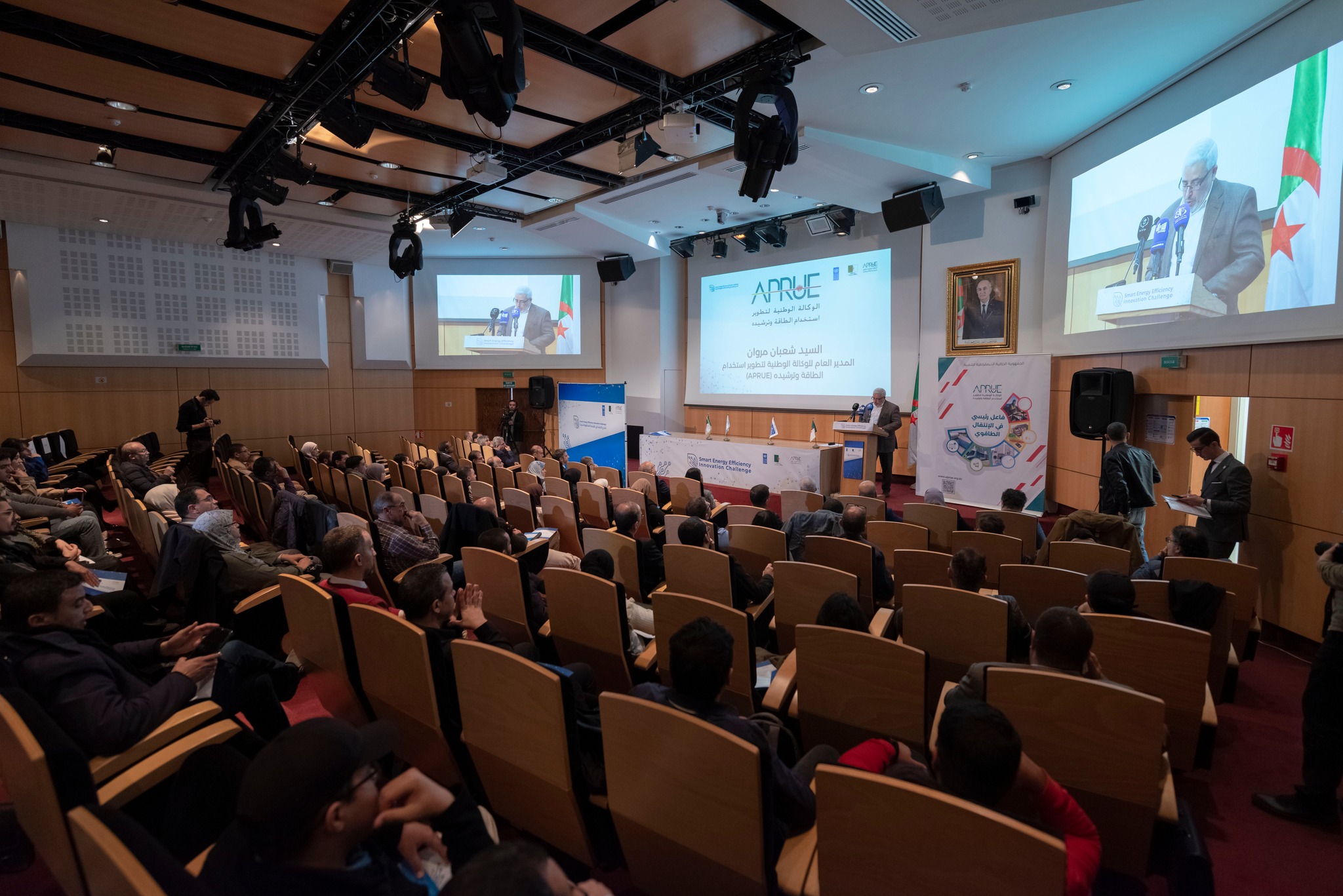 Photo: Conference hall audience watching a presenter at a podium with slides.