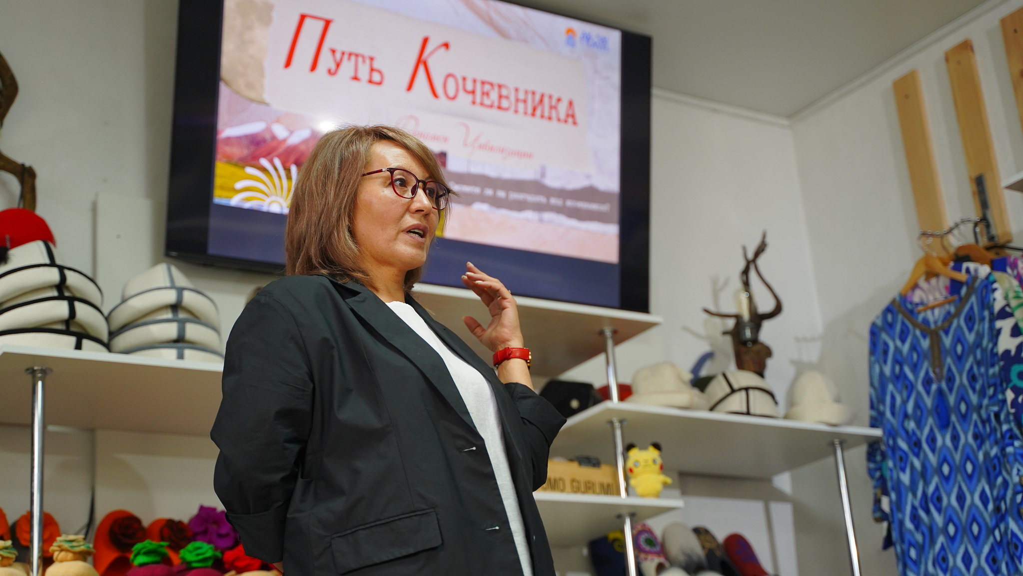 Speaker in blazer with microphone in a craft shop, shelves of hats and yarn behind.
