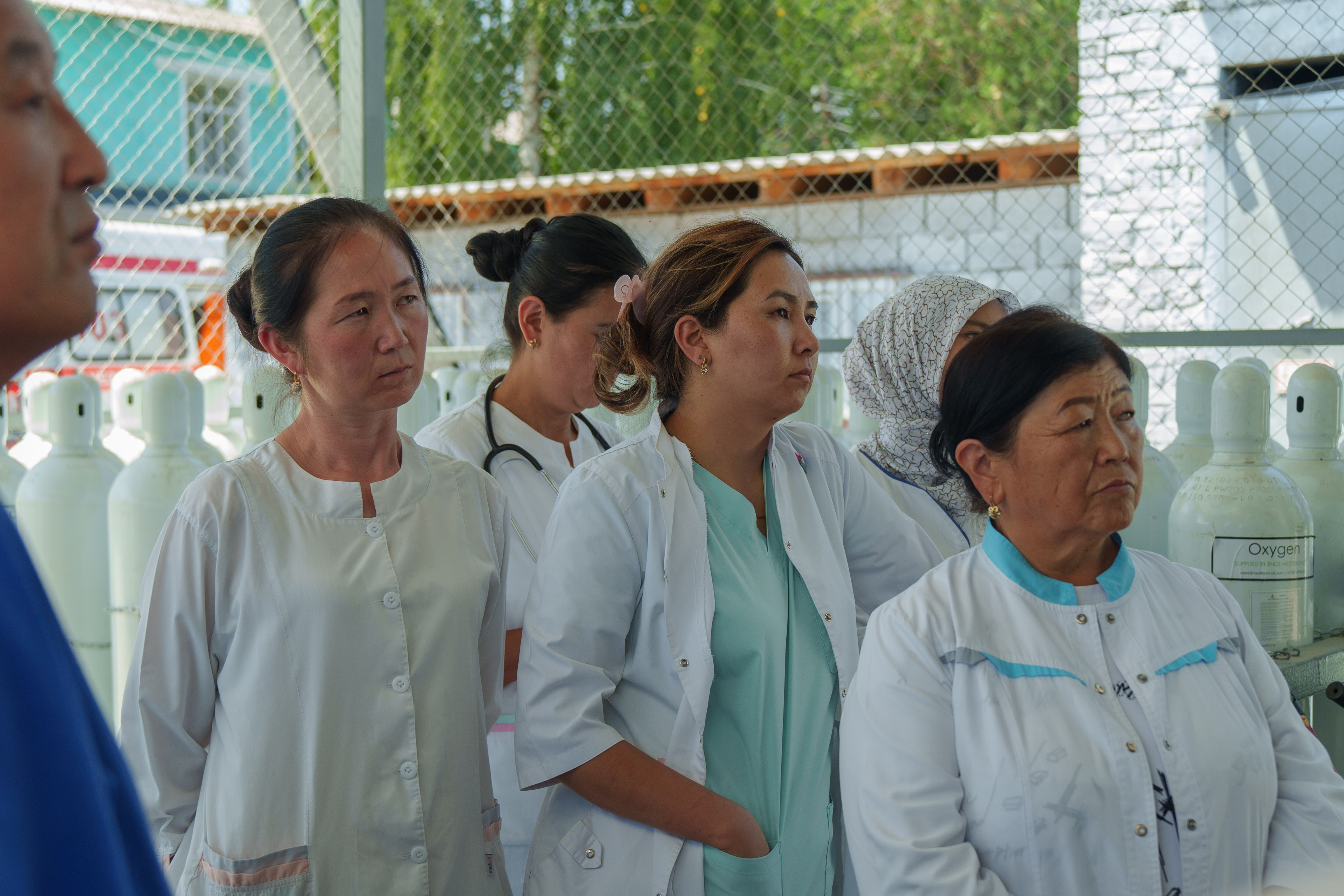Group of people in white lab coats standing outdoors beside a chain-link fence.