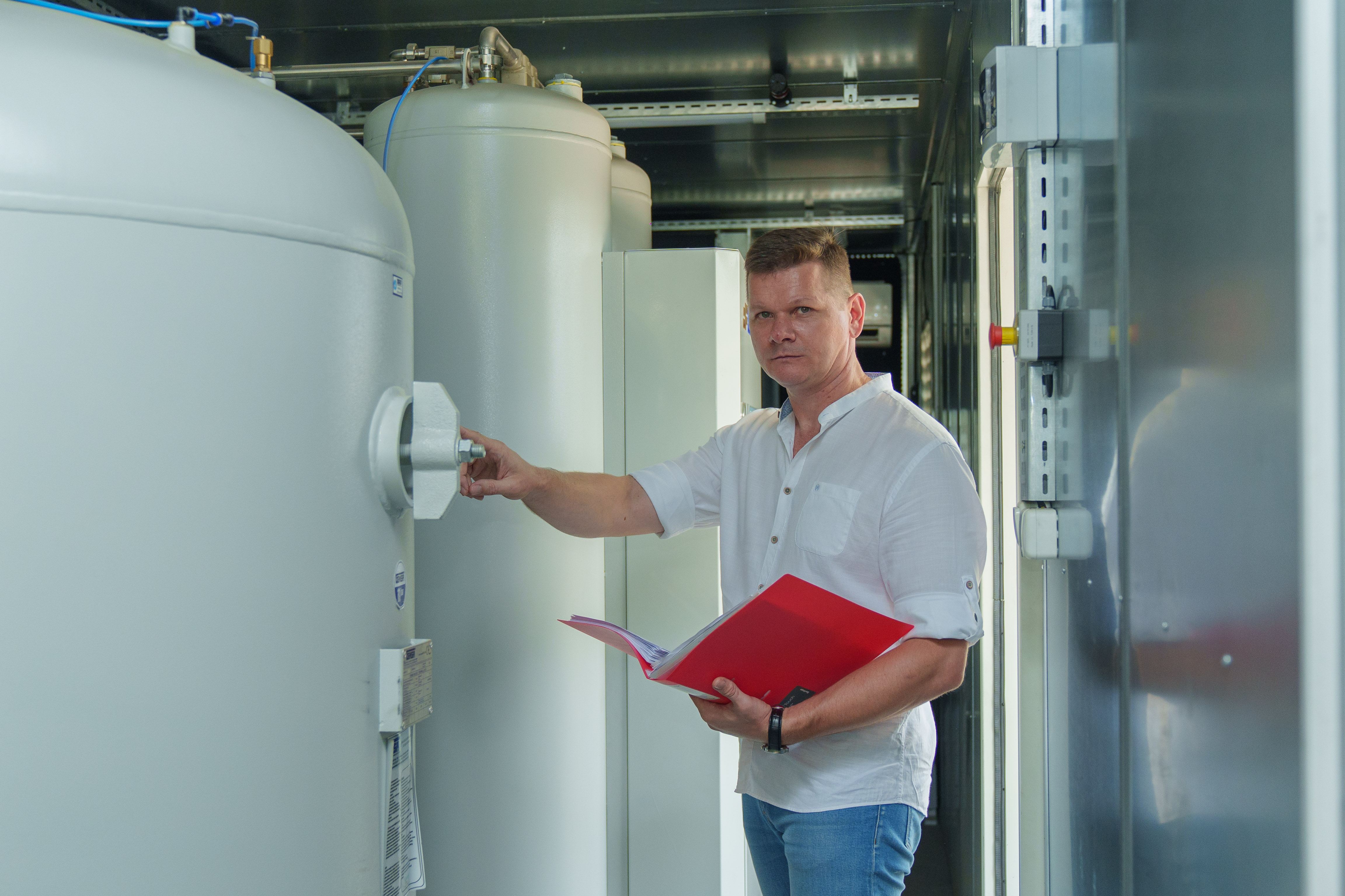 Man in a white shirt holds a red folder while inspecting large industrial tanks.