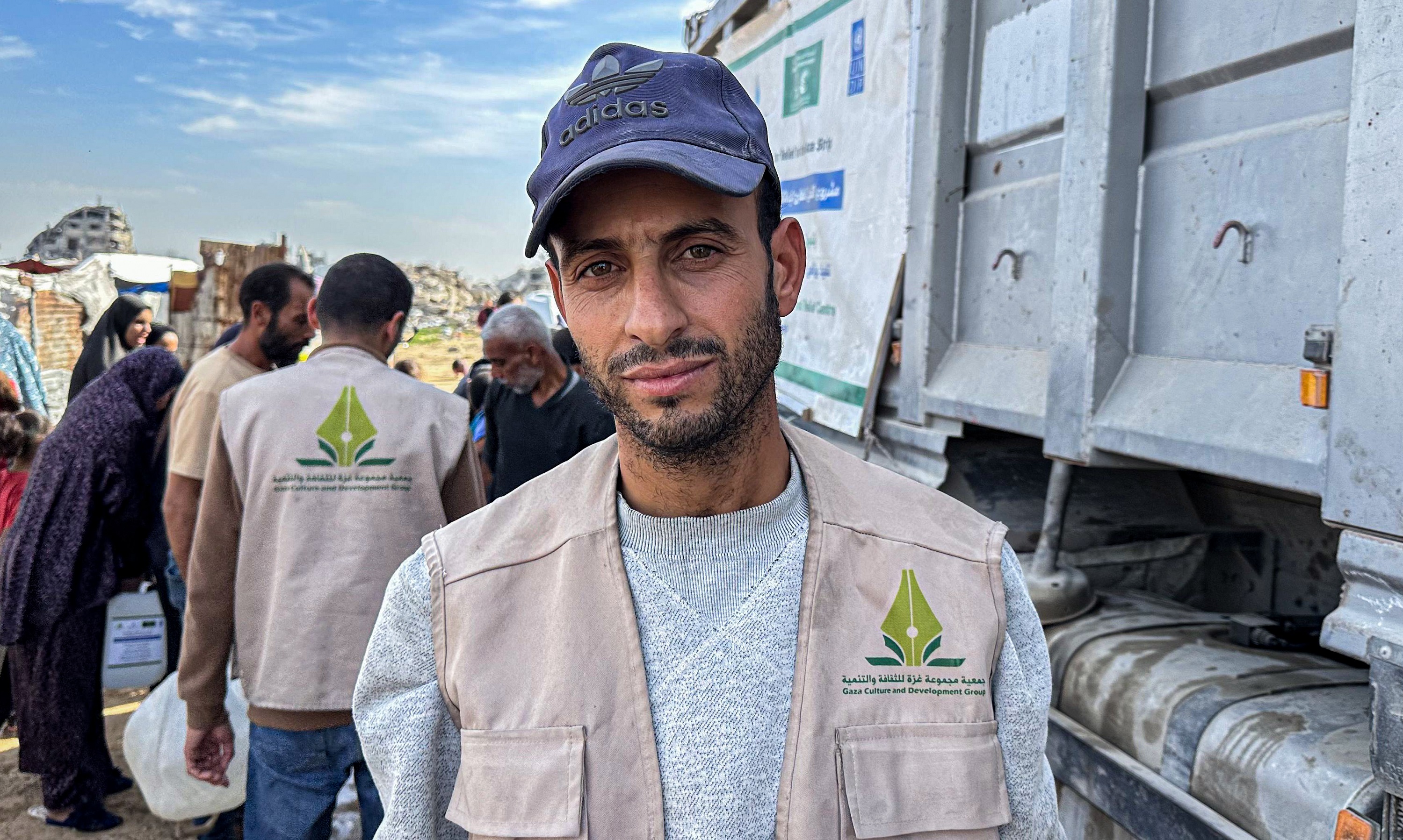 Volunteer in beige vest with logo beside a large truck, crowd in background.