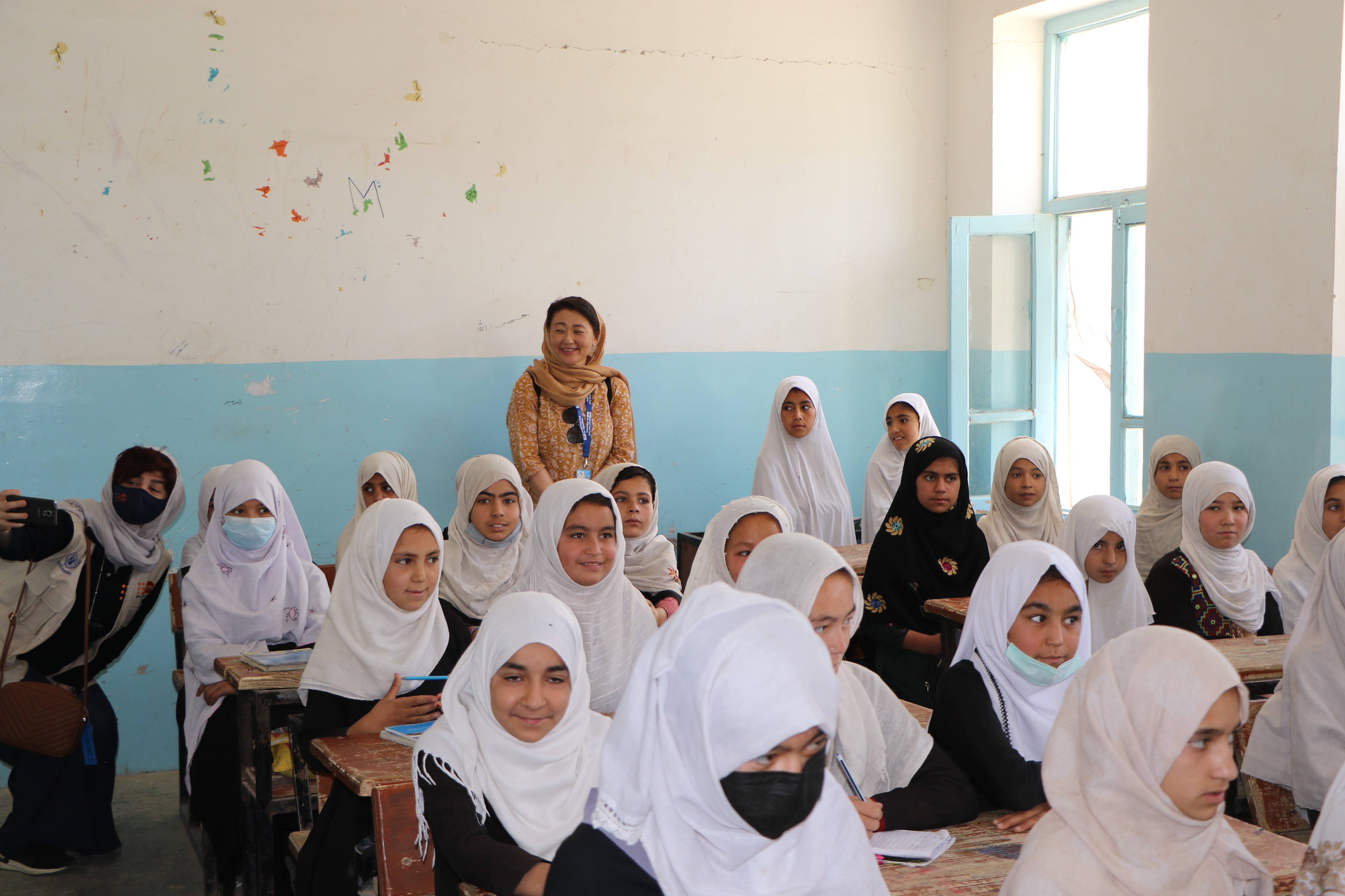 Group of students in white hijabs seated in a blue-walled classroom; a teacher stands at the back.