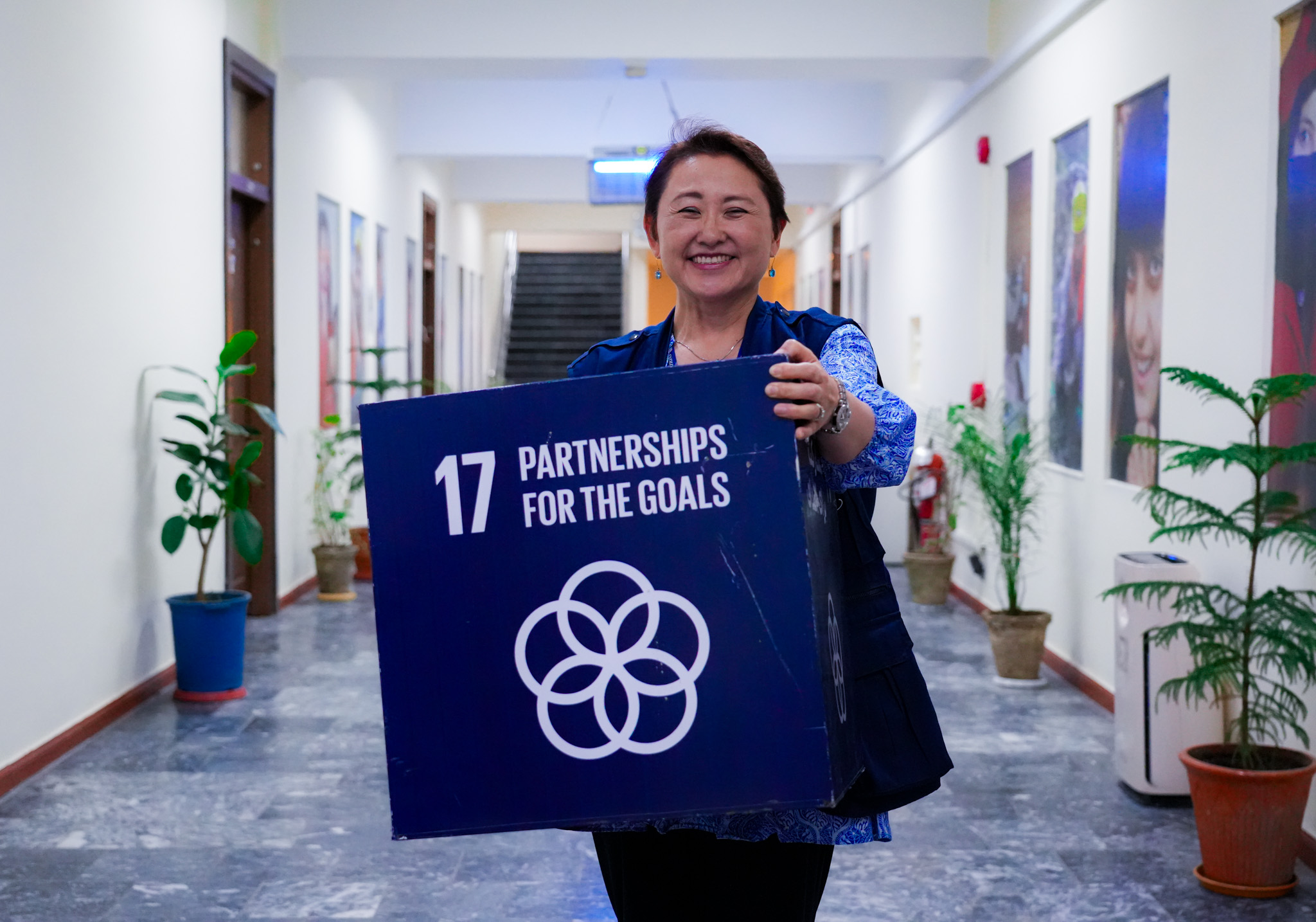 Photograph of a person in blue holding a sign that reads PARTNERSHIPS FOR THE GOALS in a hallway.