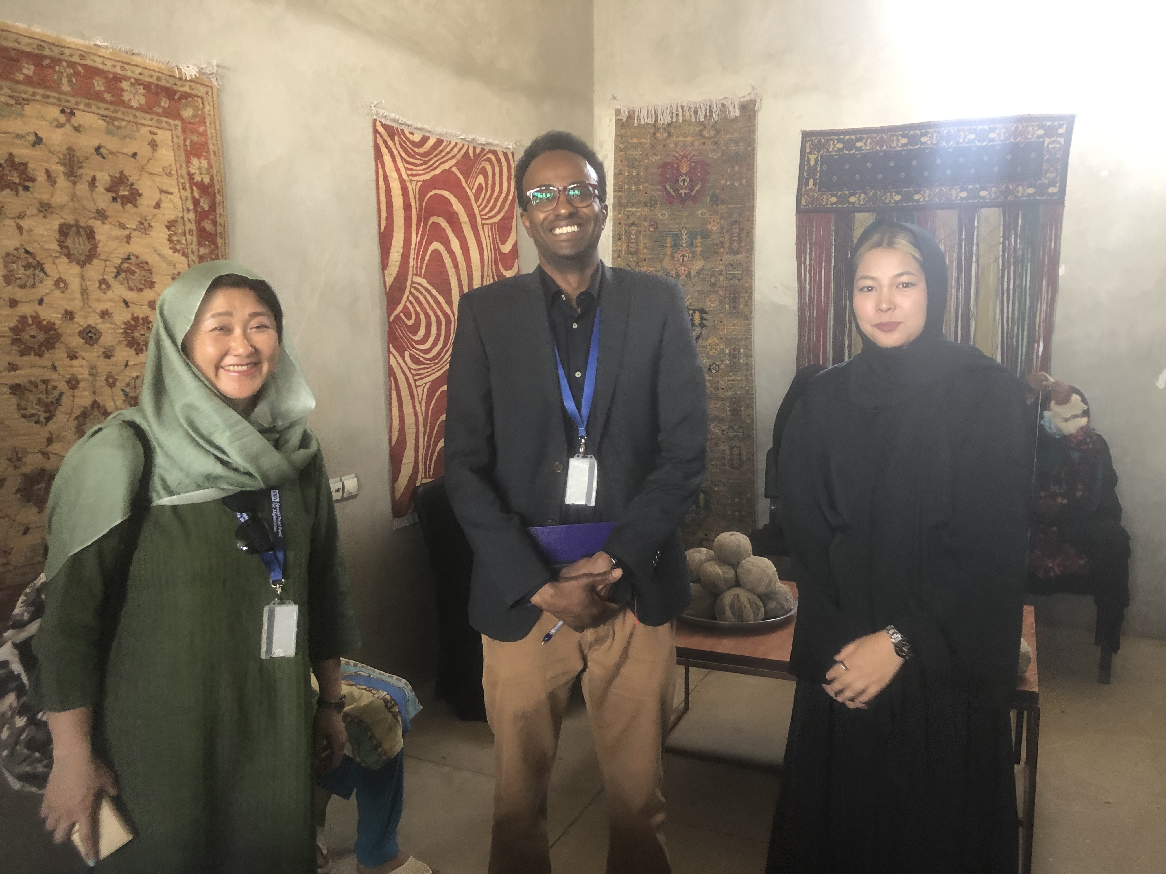 Three people indoors before rugs: left in green hijab, center with glasses, right in black abaya.