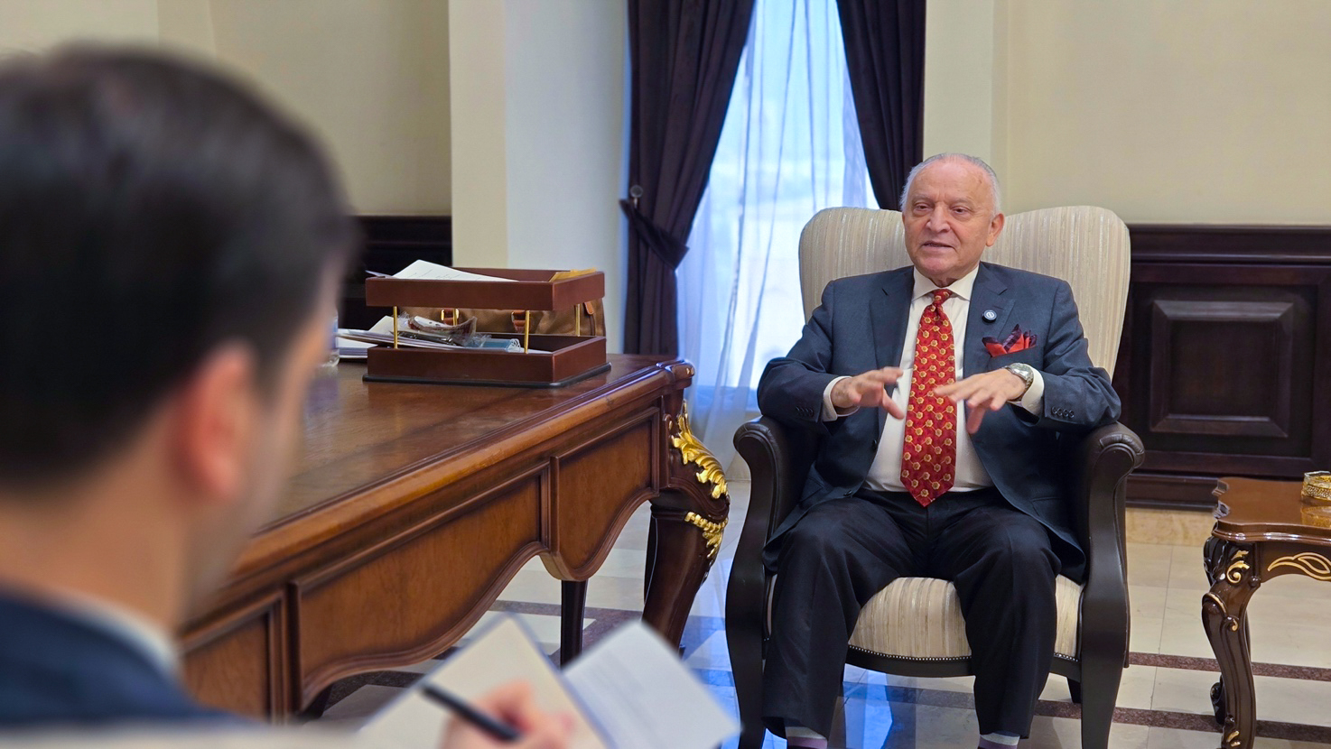 Photograph: two men in a wood-paneled office; one gestures while the other writes.
