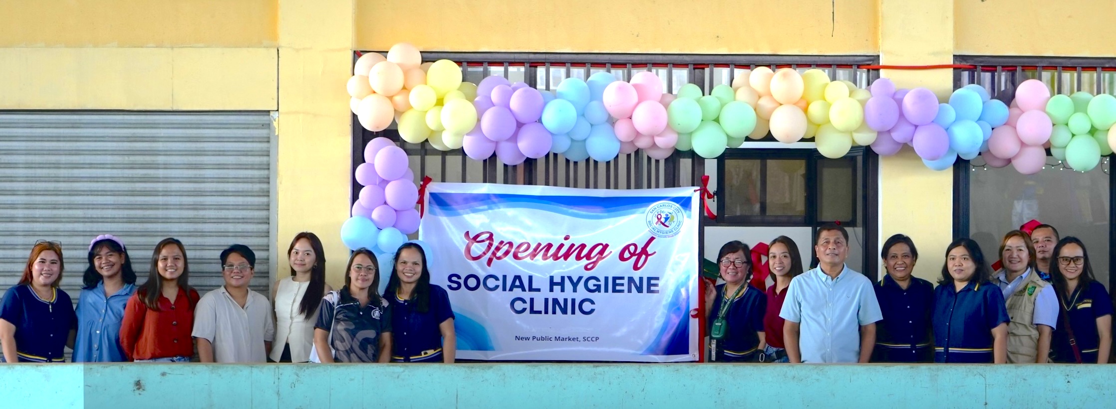 Photo of a group on a balcony with colorful balloons and a banner in the background.