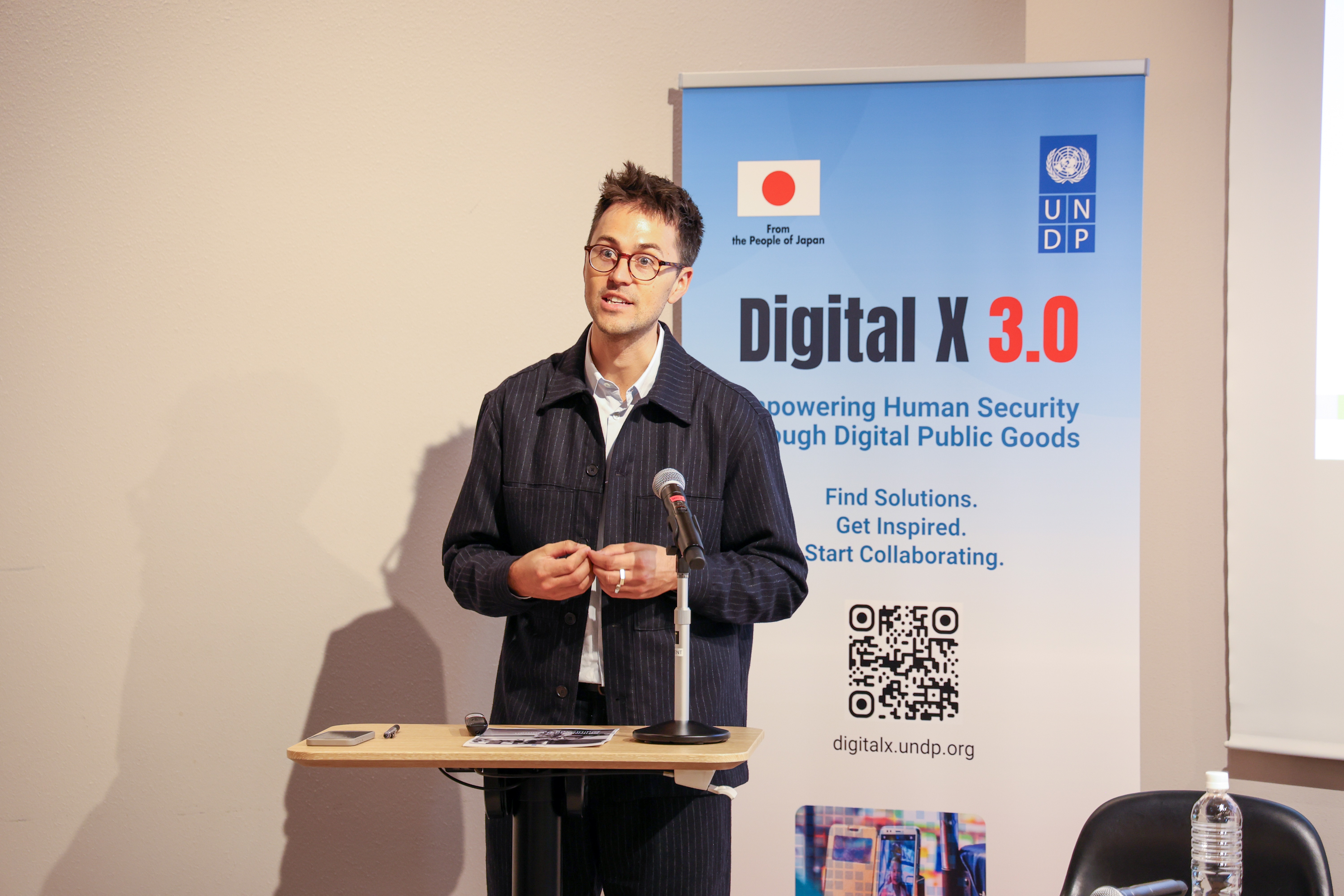 Male speaker with glasses at a podium in a conference room, banner behind reads Digital X 3.0.