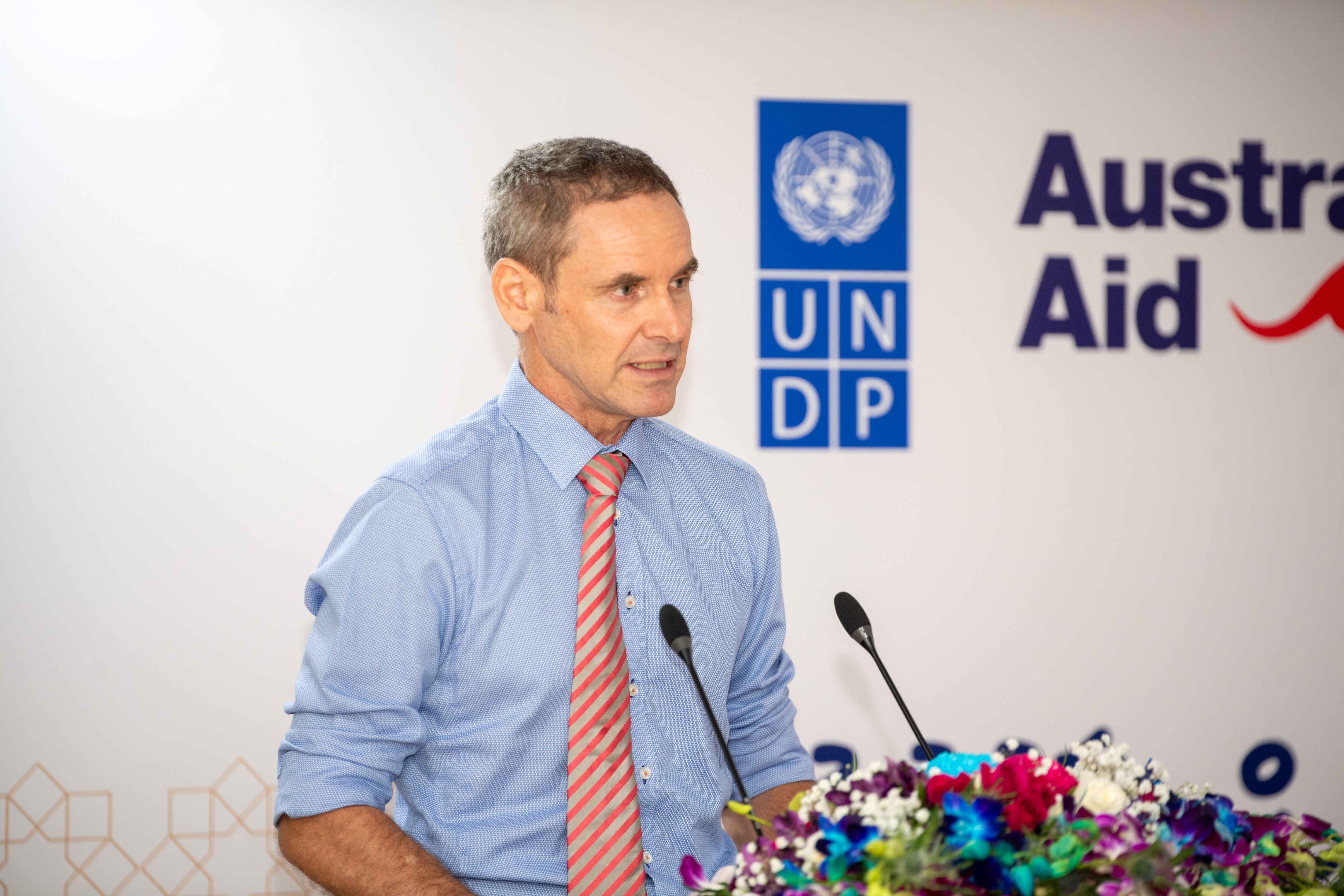 Speaker in blue shirt and pink striped tie at podium with UNDP and Australian aid banners, flowers.