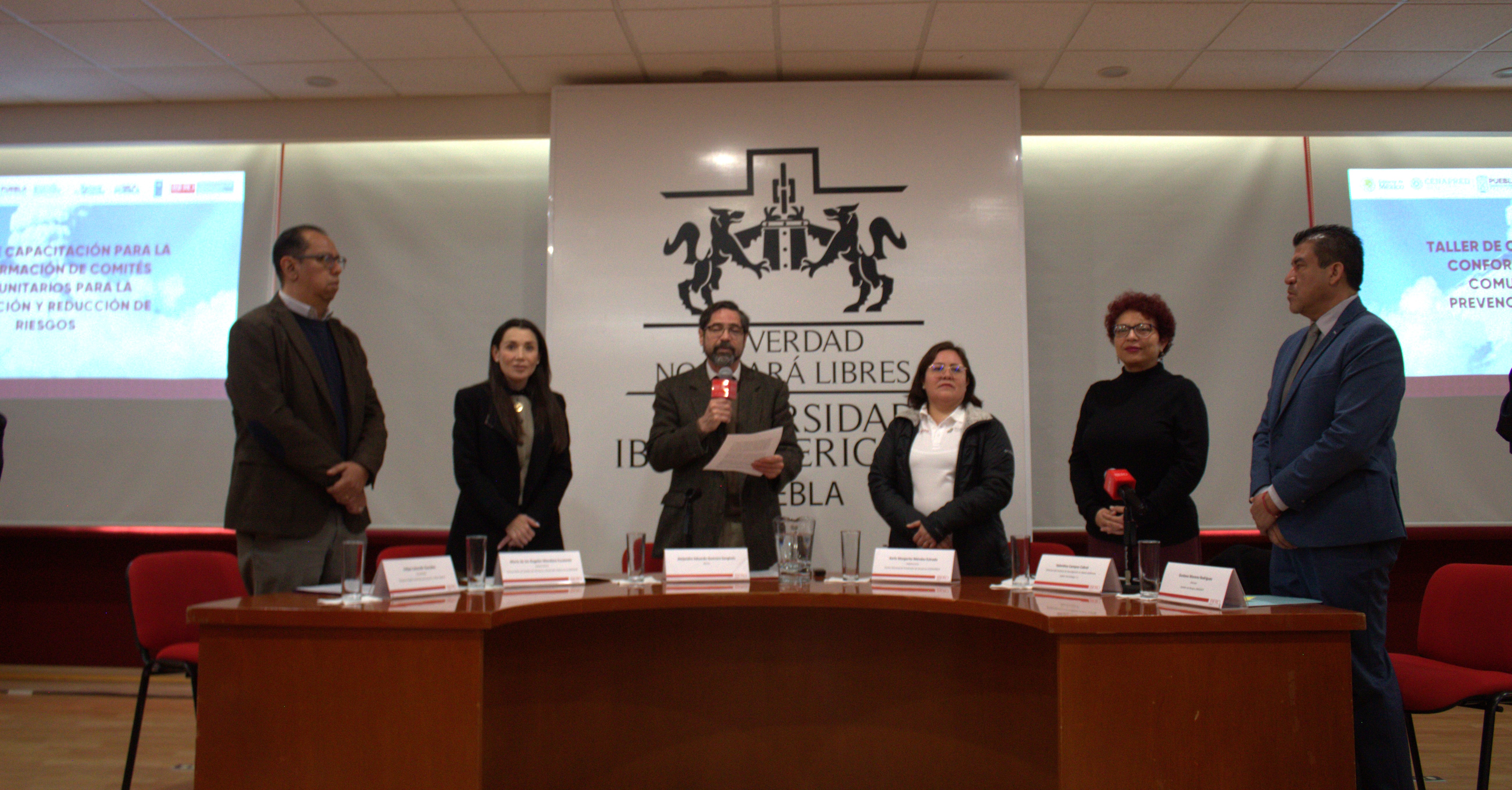 Group of people standing behind a long conference table on a stage, crest banner in the background.