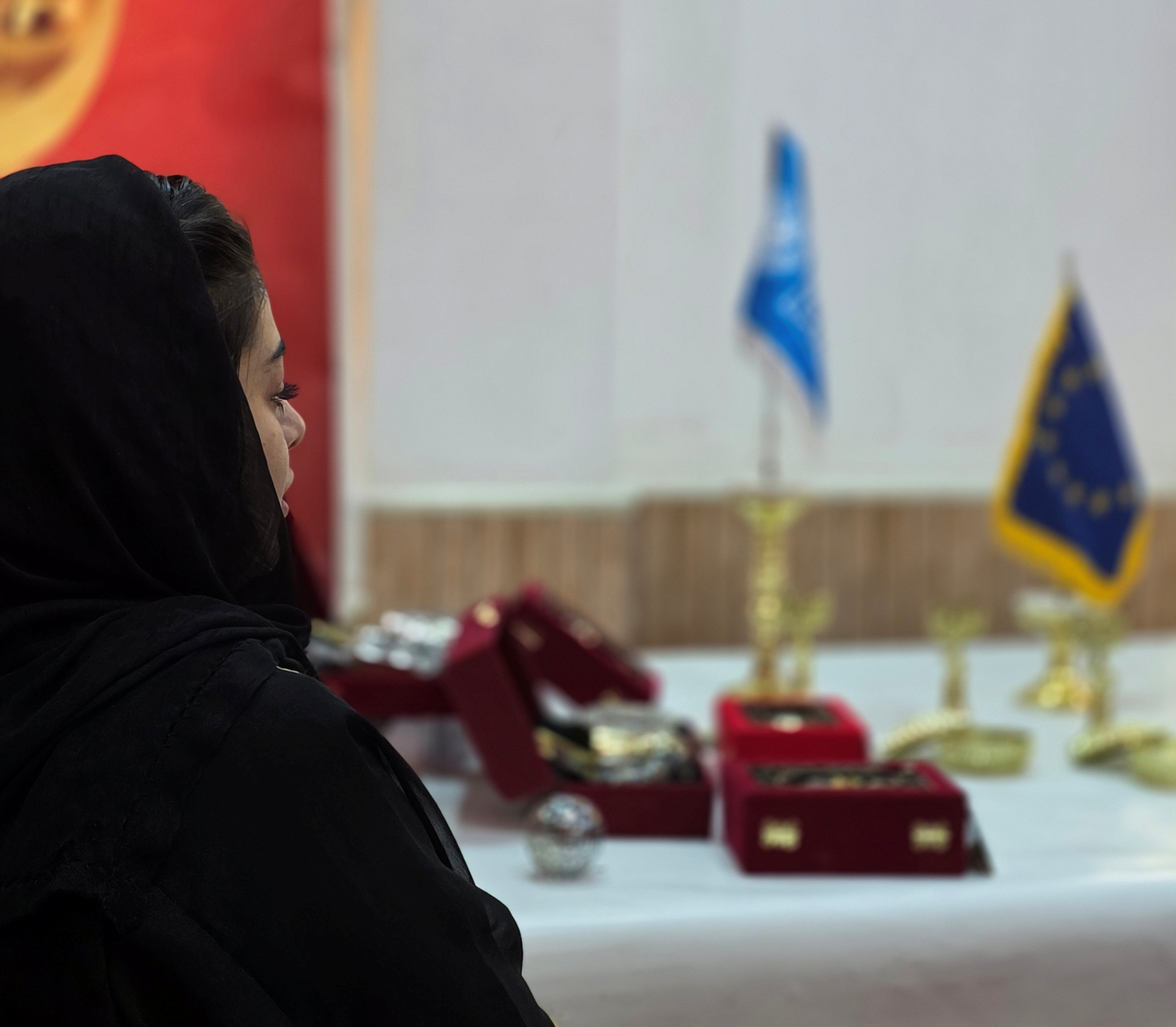 Woman in a black hijab examines a table display of red jewelry boxes; flags behind.