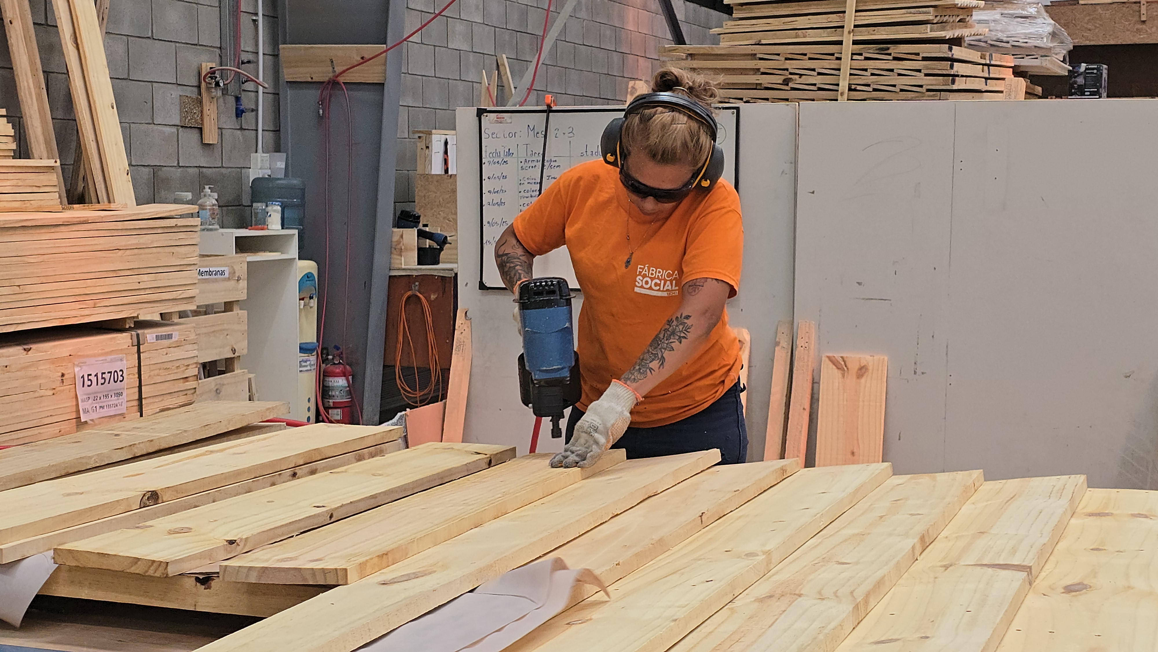 Construction worker in orange shirt and helmet cuts wood planks with a power saw at a lumberyard.