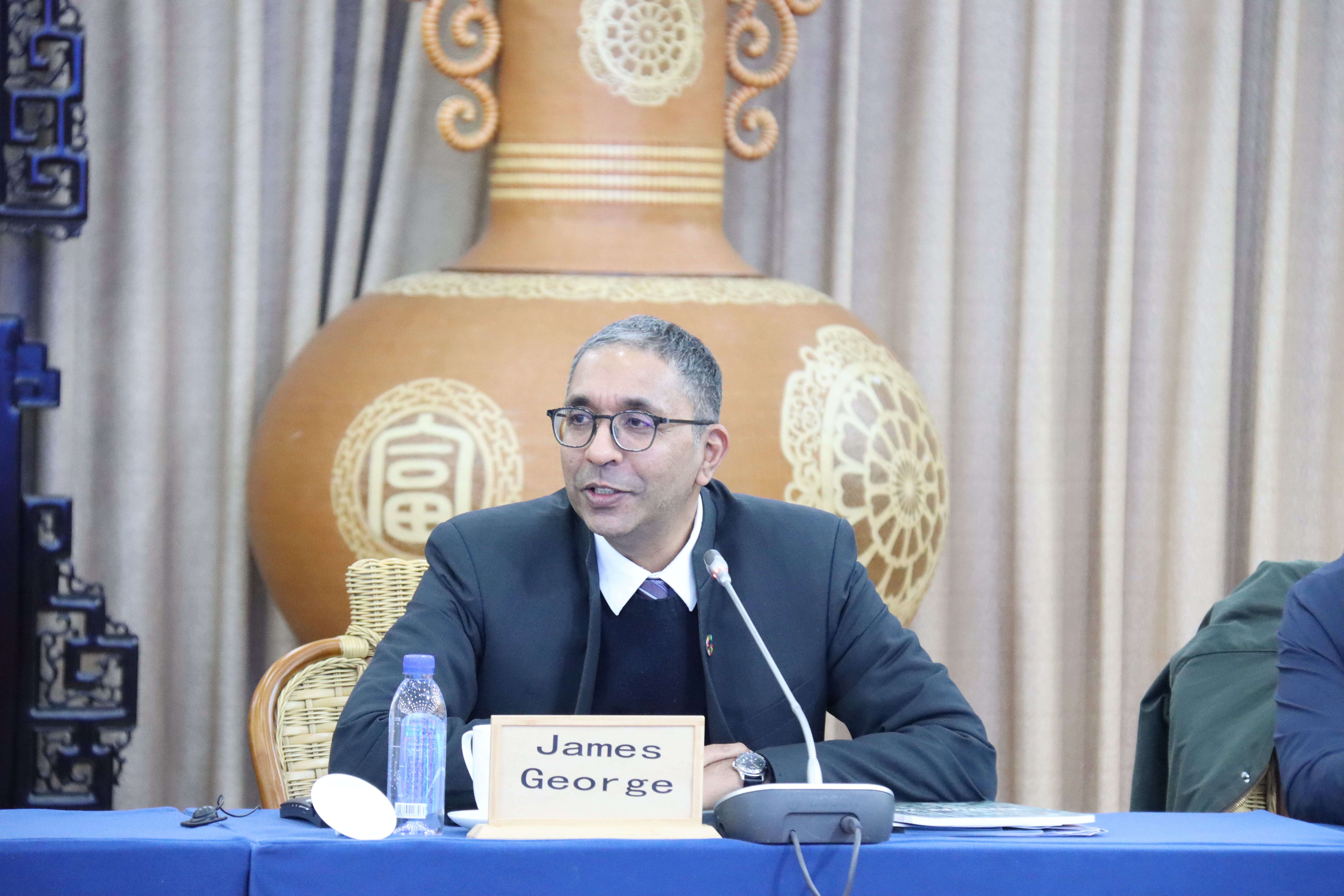 Conference panel with a suited man, blue table, nameplate James George, ornate vase backdrop.