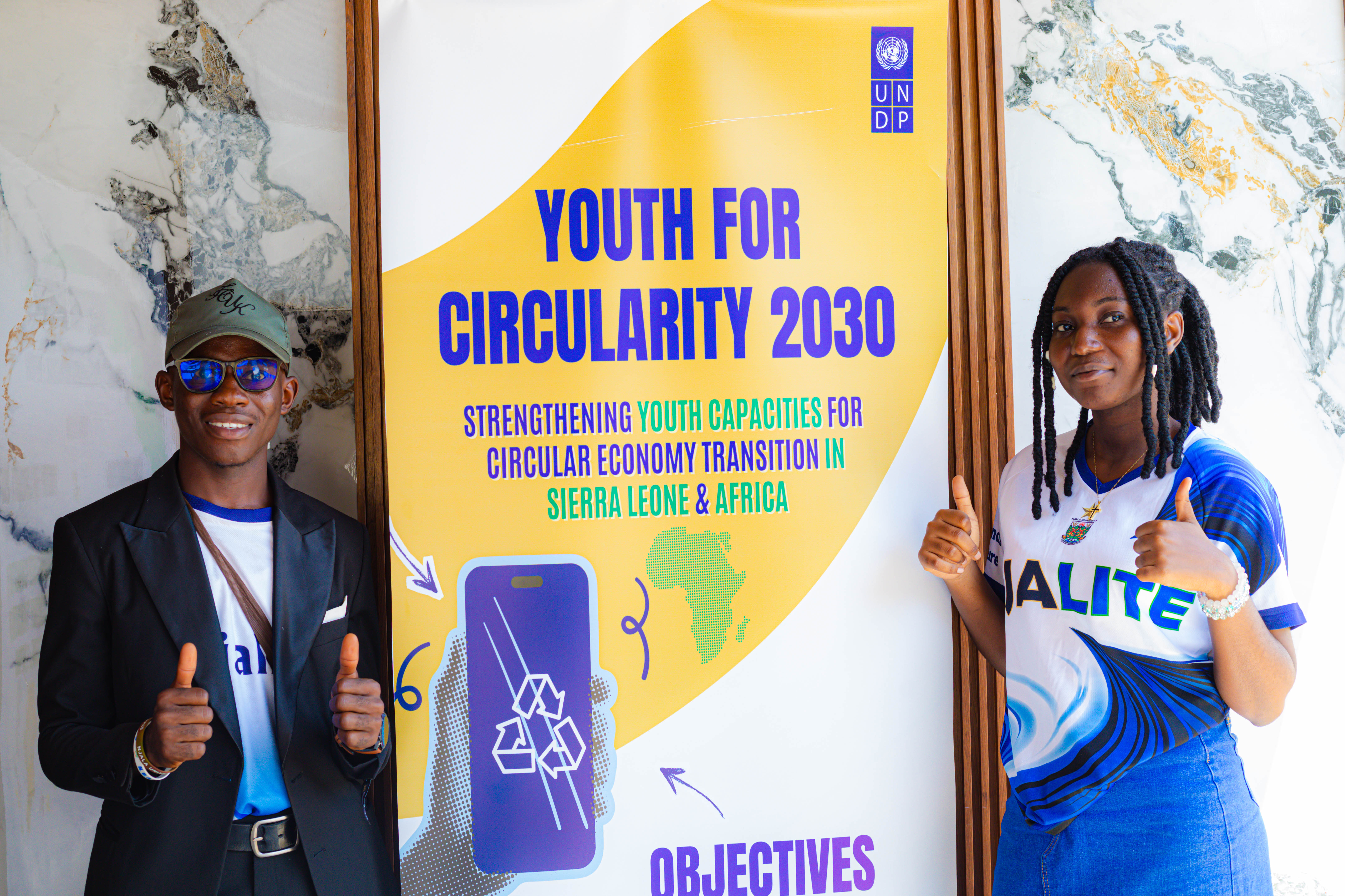 Two people beside a yellow banner reading 'Youth for Circularity' with a recycling symbol.
