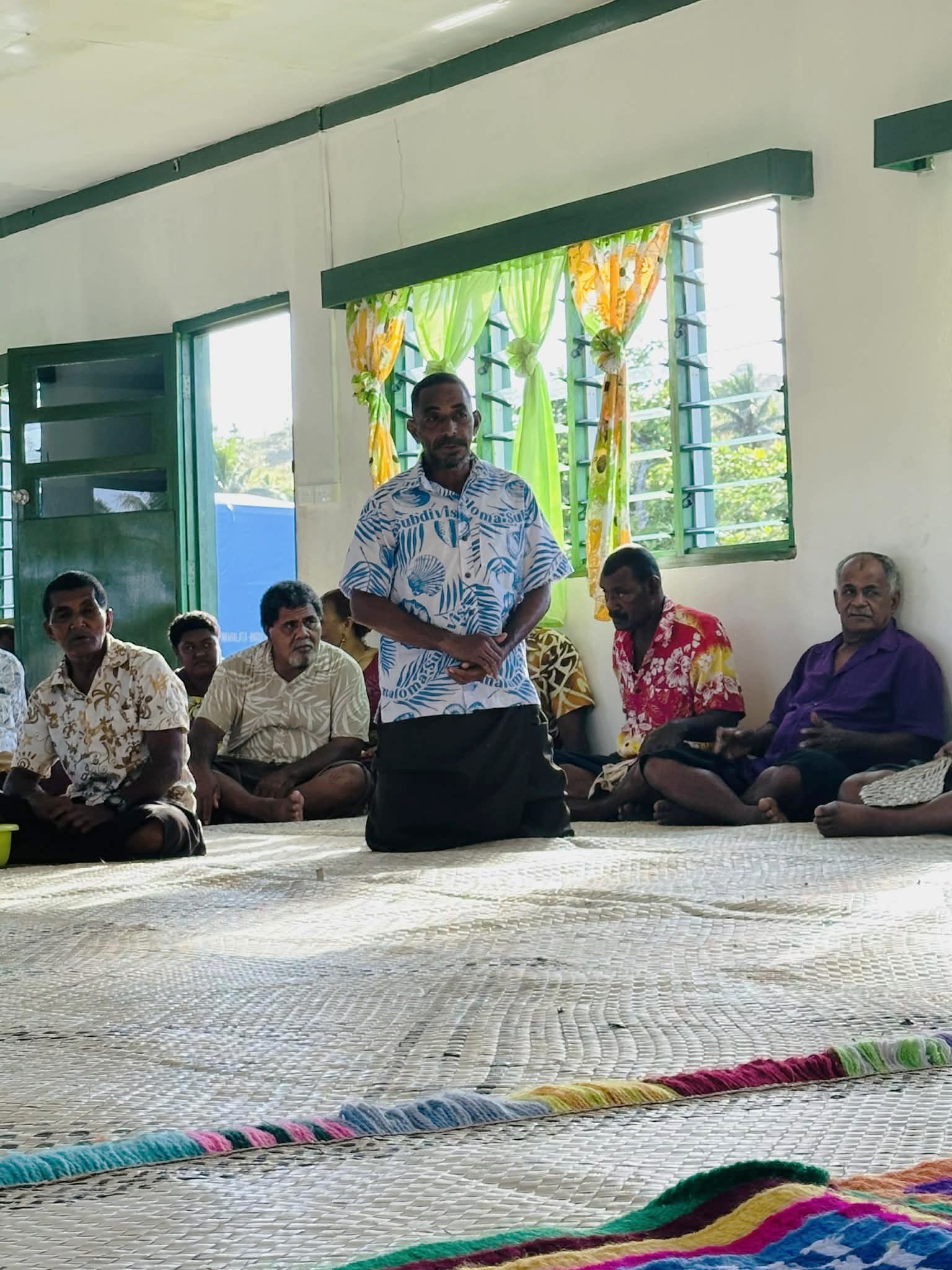 Man stands and speaks to a seated group on a woven mat in a bright room.