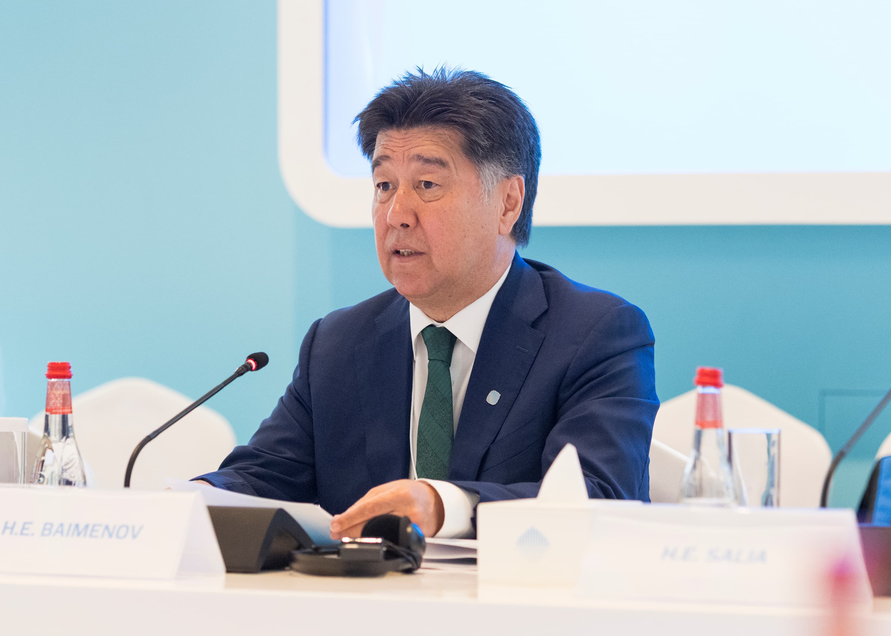 Speaker in a suit and green tie sits at a conference table with microphones, water, blue backdrop.