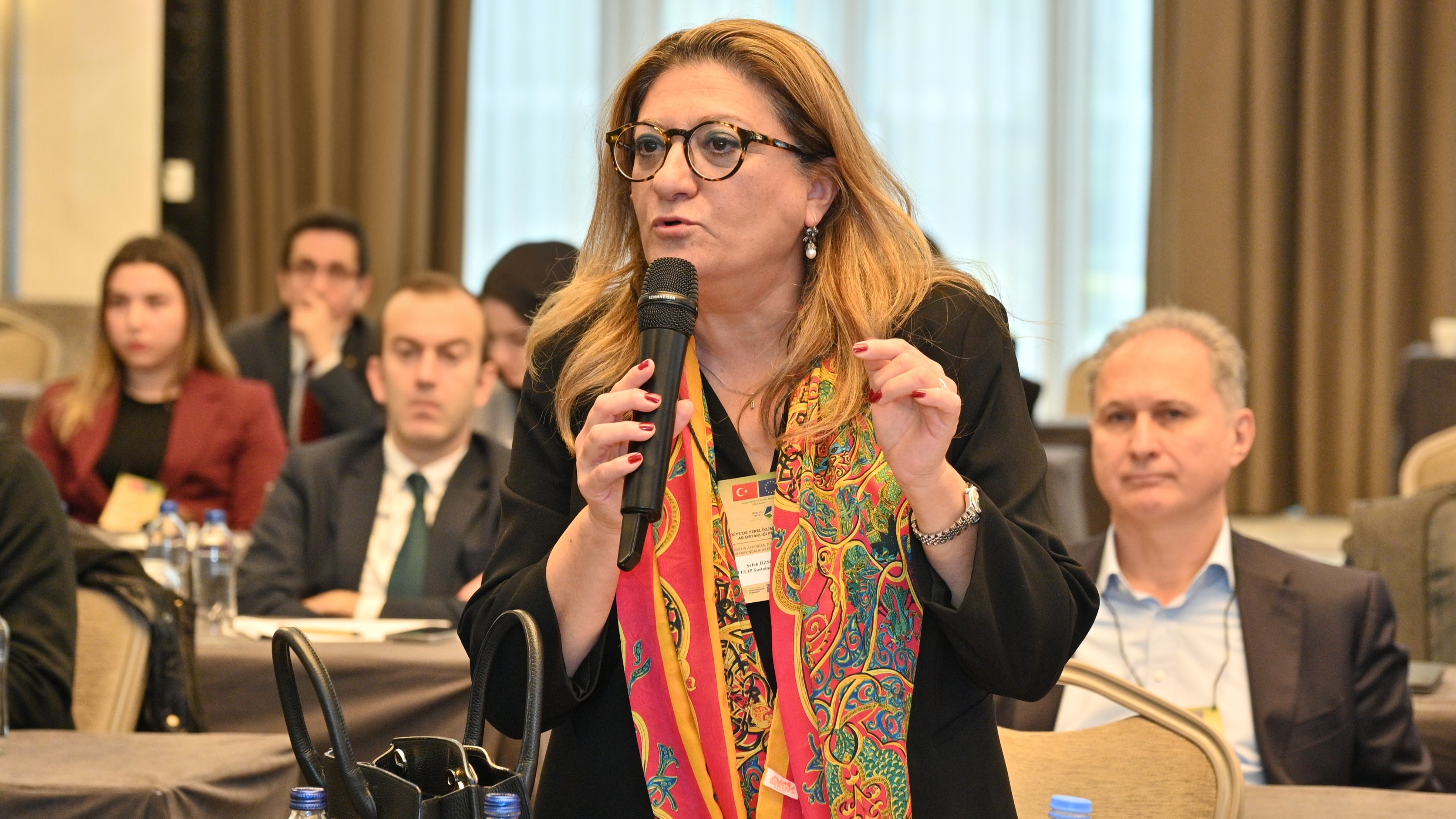 Speaker with a colorful scarf holding a microphone at a formal conference.