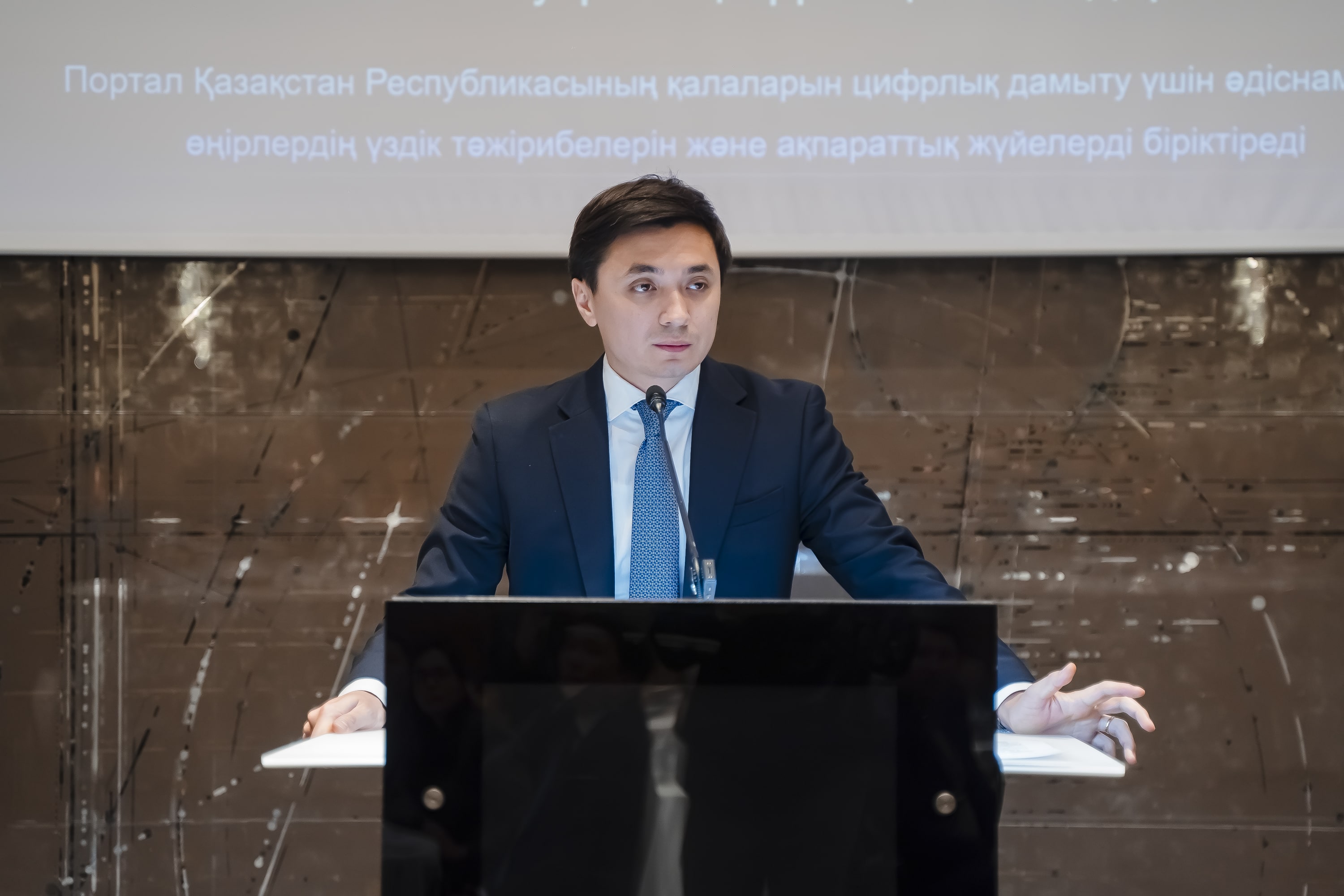 Speaker in suit at a black podium, arms on the sides, backdrop features marble wall and banner.