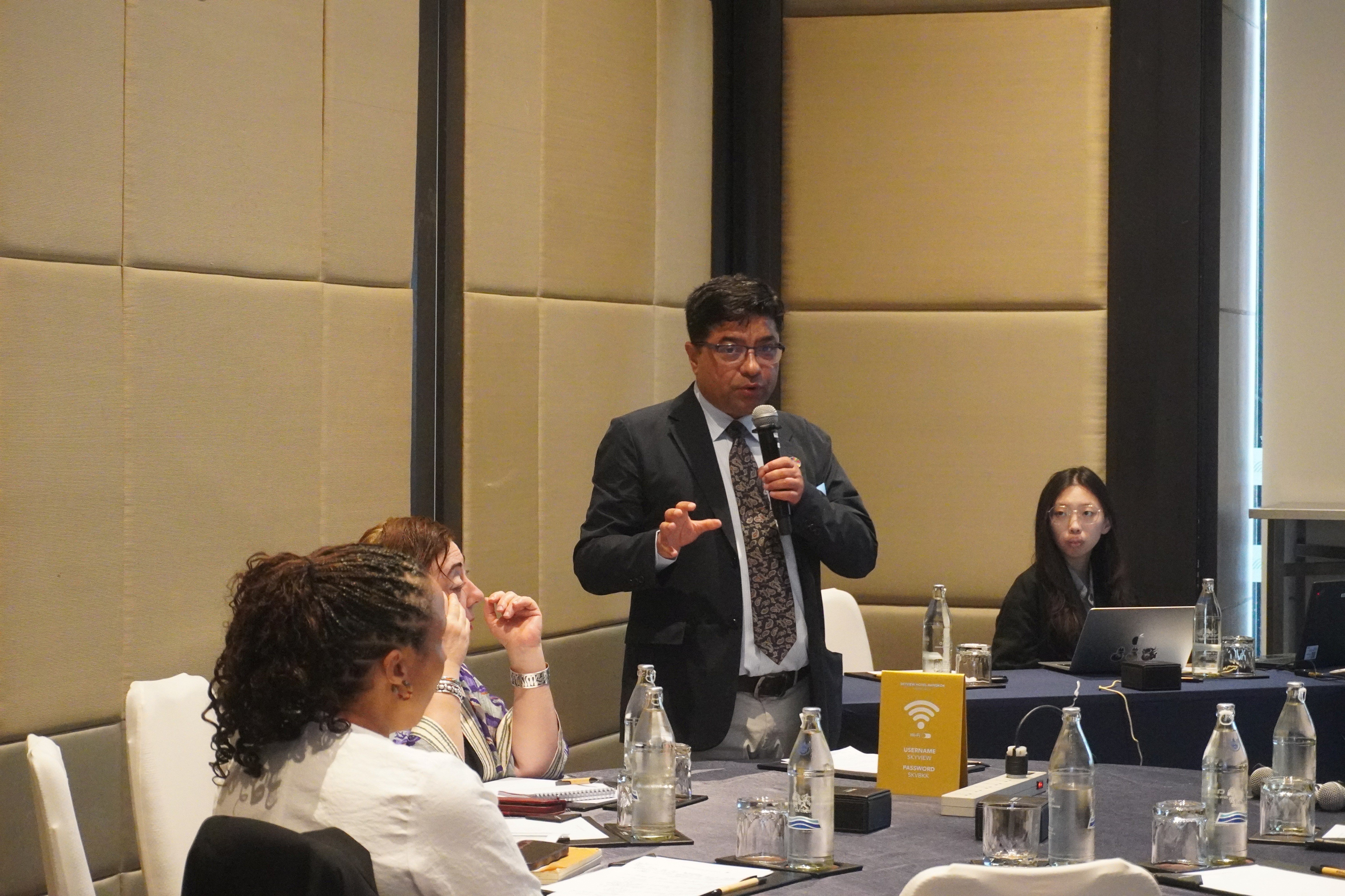 Man in suit speaks into microphone at panel discussion, with seated attendees and water on table.