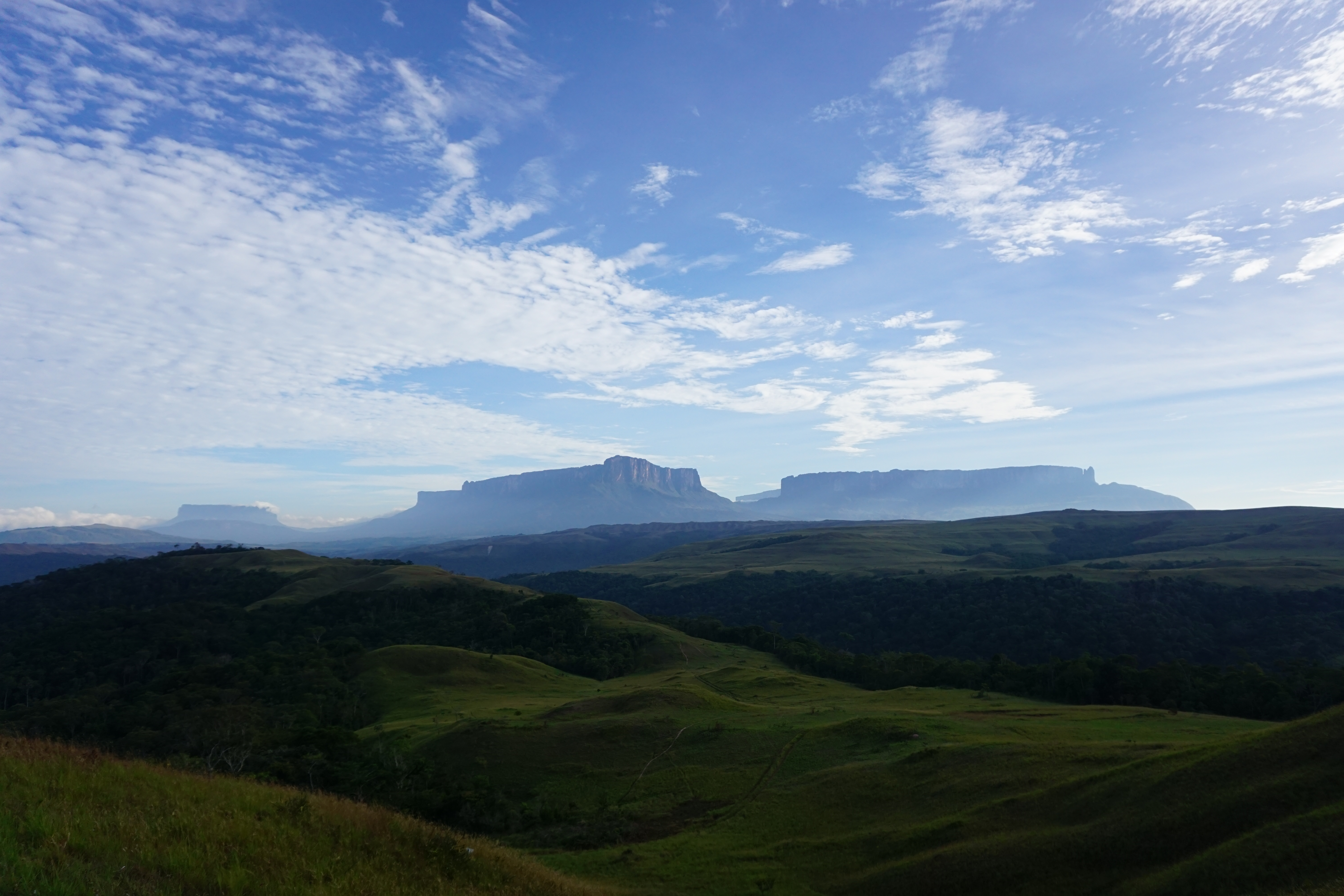 Wide view of green rolling hills with distant flat-topped mesas under a blue, cloud-dappled sky.