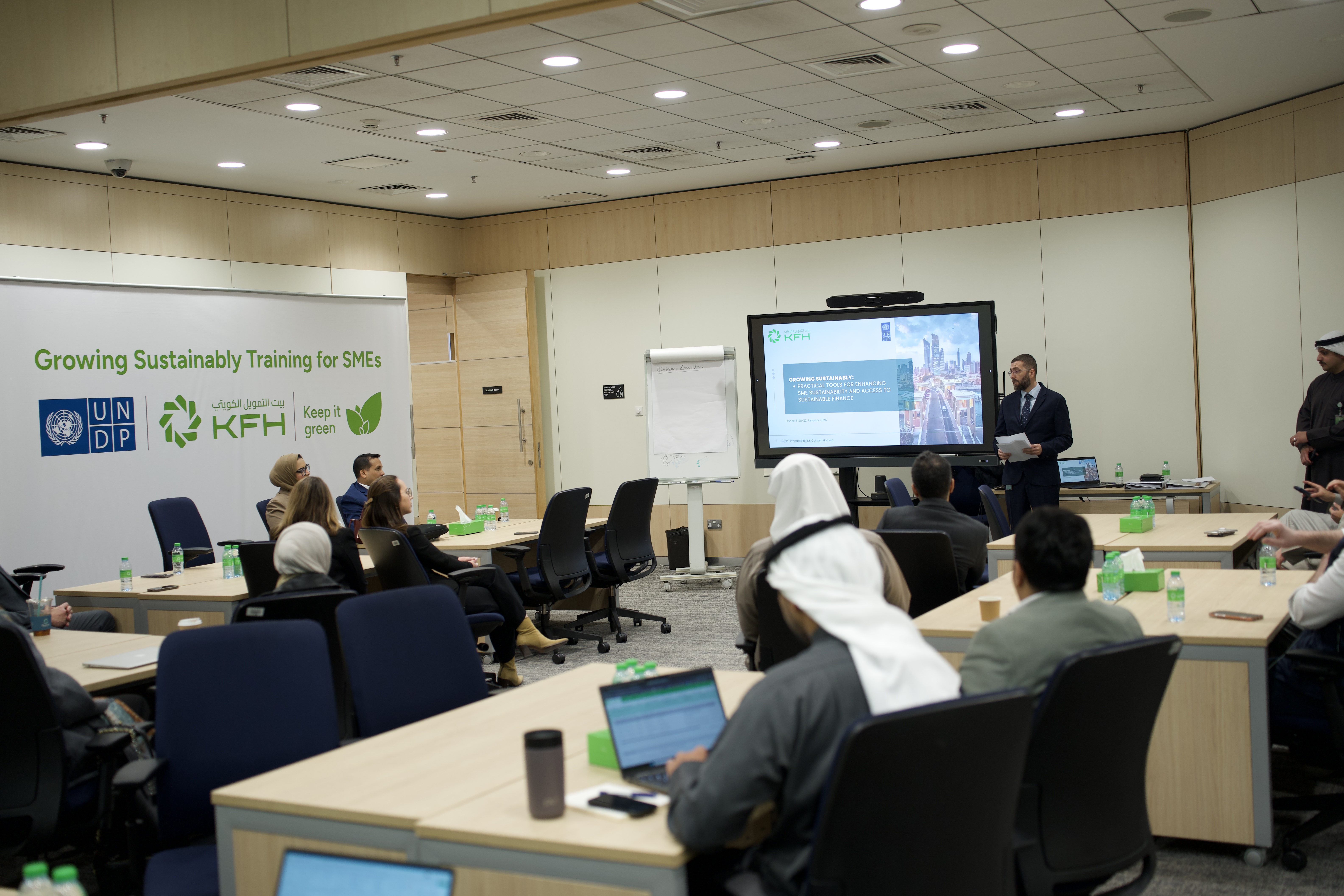 Workshop room with attendees at tables, laptops open, and a presenter by a large screen.