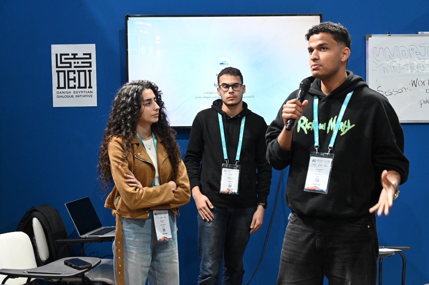 Three people with lanyards in a blue room; the right person speaks into a microphone.