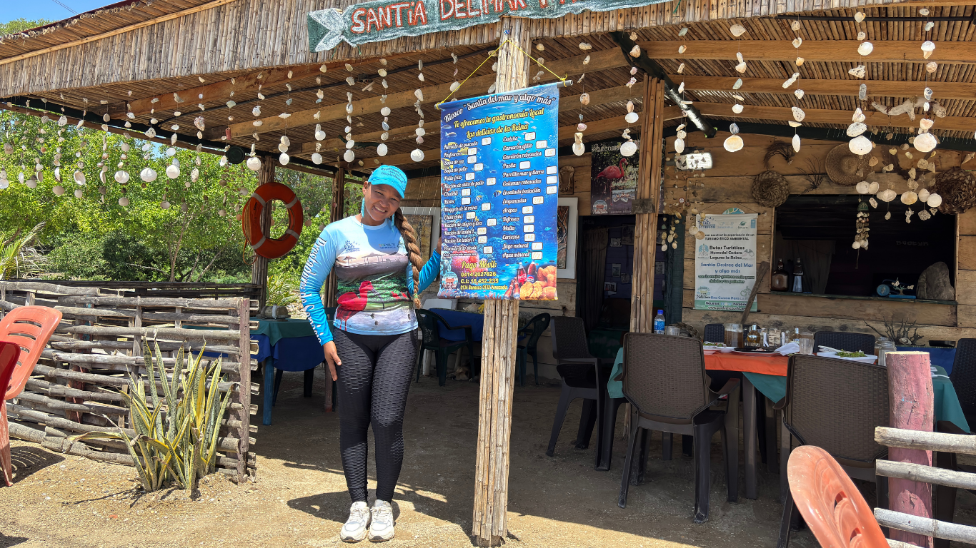 Person in a blue shirt near a thatched-roof beachside cafe with string lights and plastic chairs.