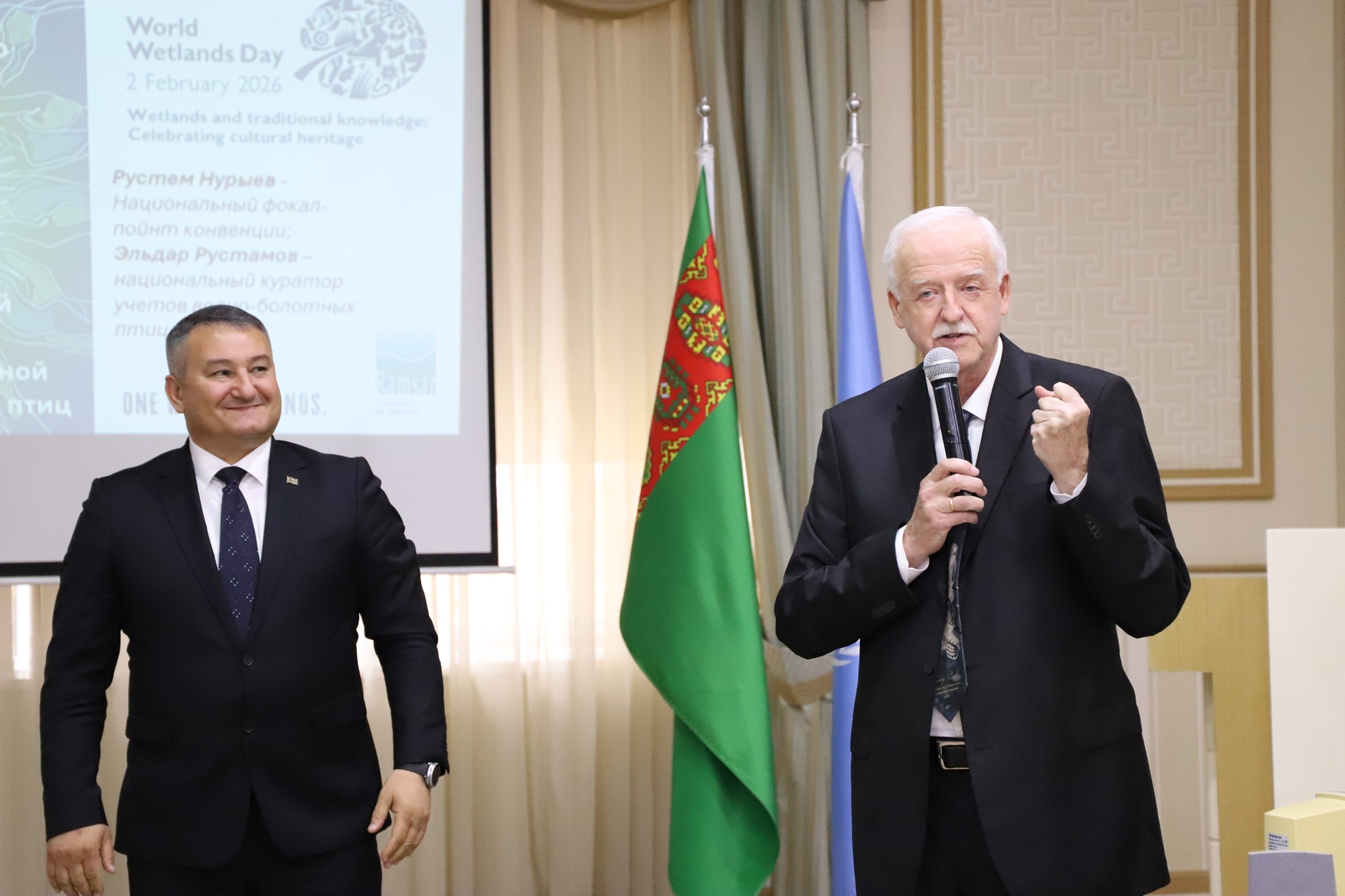 Two men in suits on stage; one speaks into a microphone, with a flag and projection screen behind.