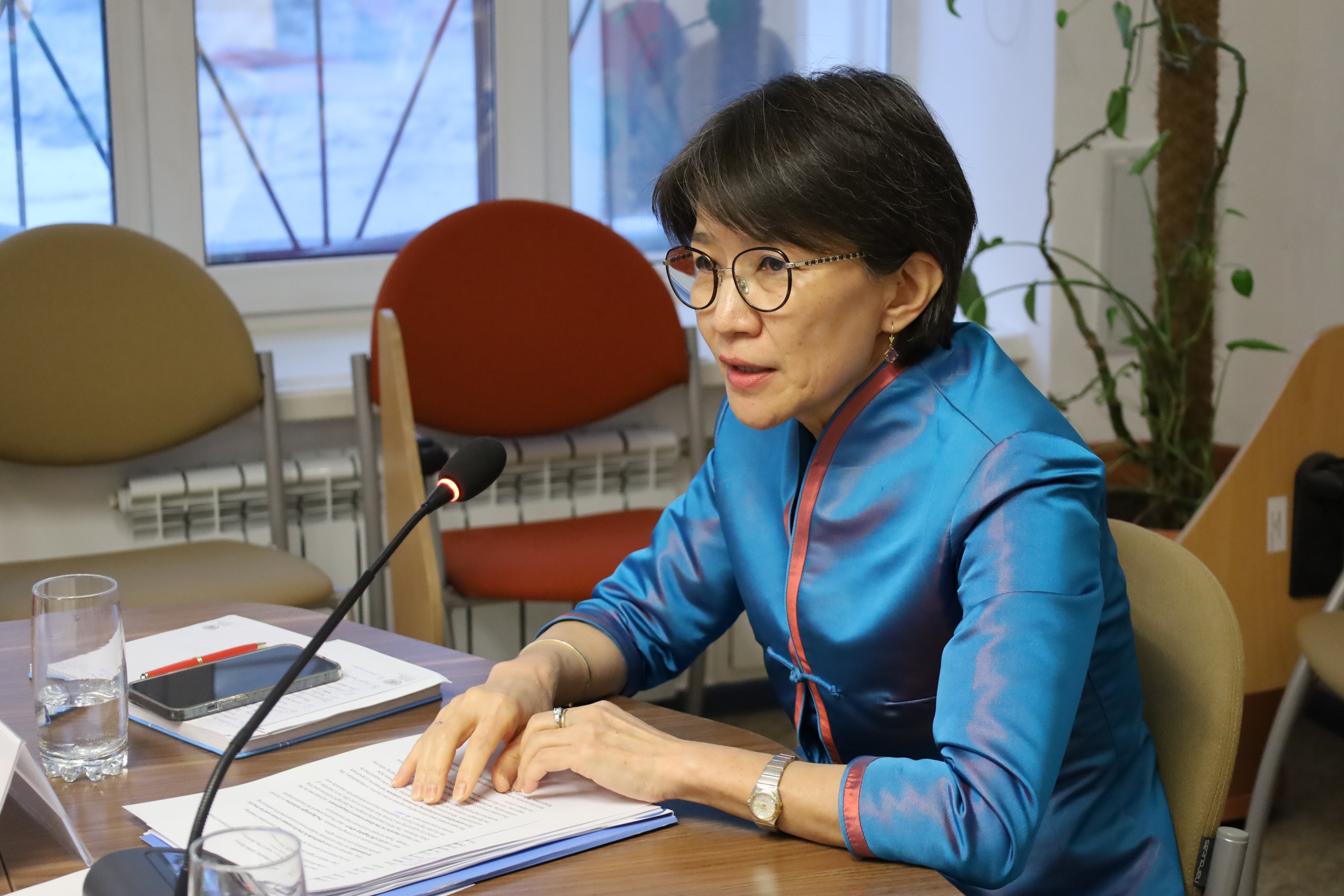 Person in blue shirt sits at a table with papers, a glass of water, and a microphone in a bright room.