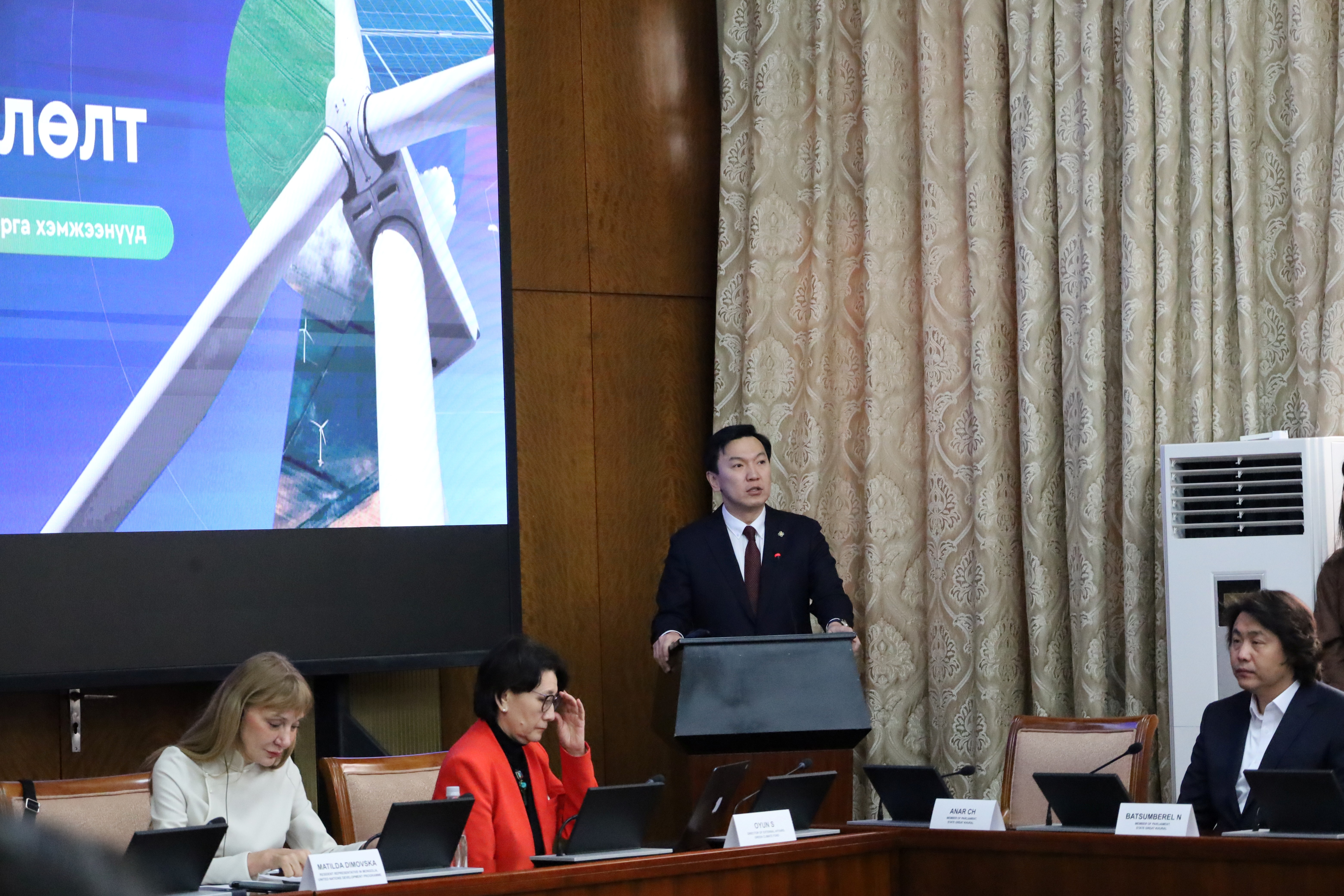 Man in suit at podium delivers talk; panelists sit at a long table, projection screen to the left.