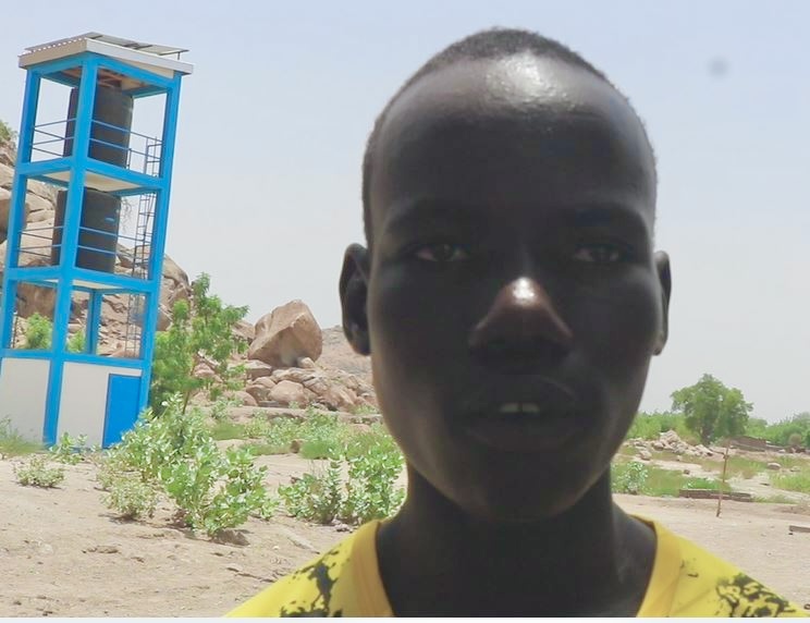 Person in yellow shirt stands outdoors in a desert-like area with a blue tower on the left.