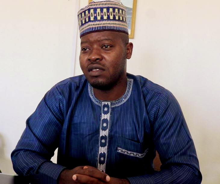 Person in blue traditional outfit and patterned cap sits with hands folded.