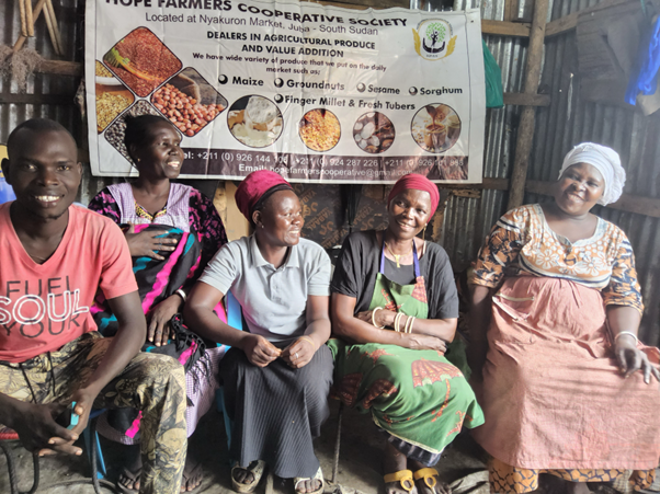 Group of women in colorful clothing sit on a bench in front of a banner at a rural community event.
