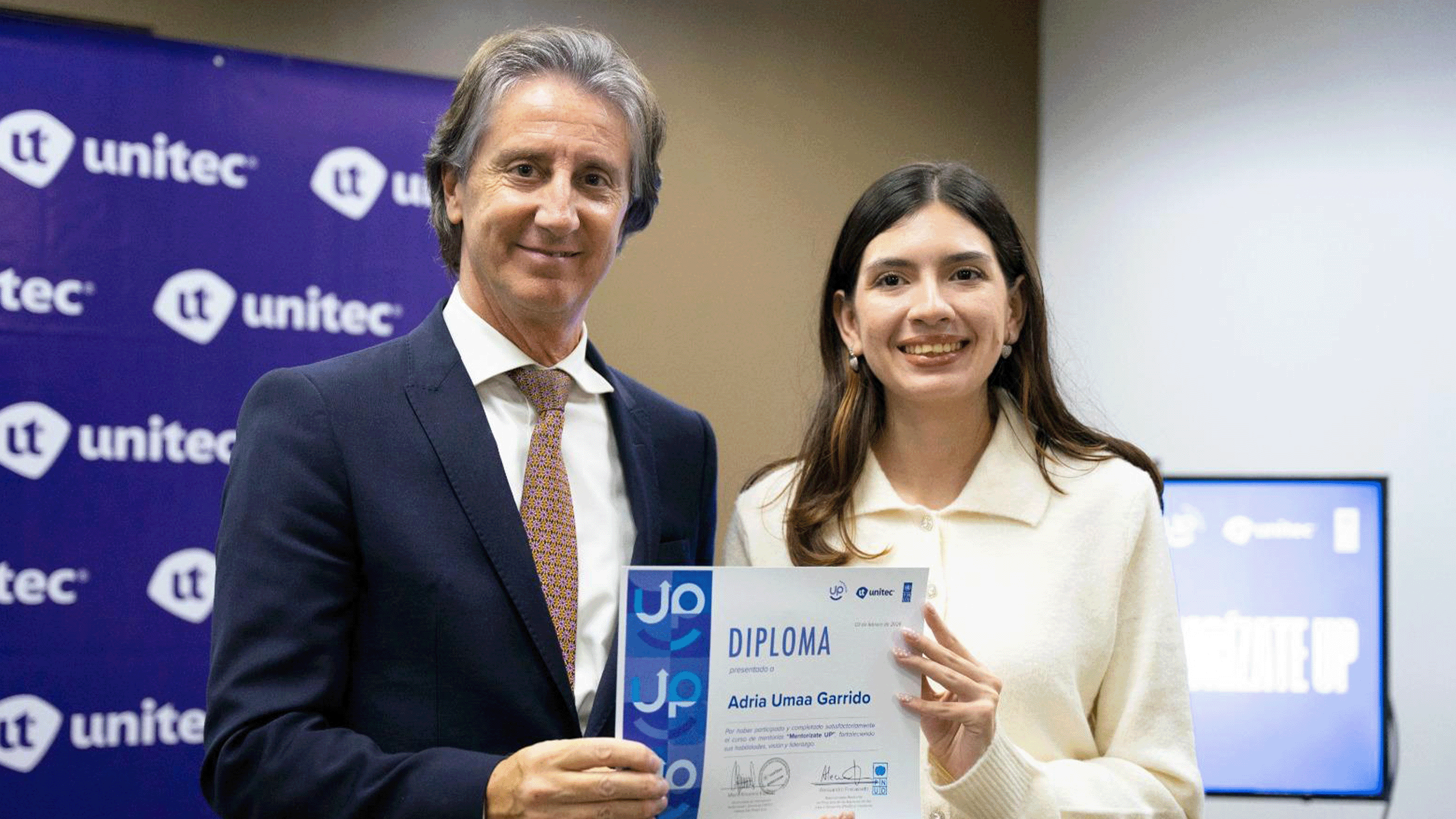 Man in suit and woman in light blazer hold a certificate at a Unitec event, blue backdrop.