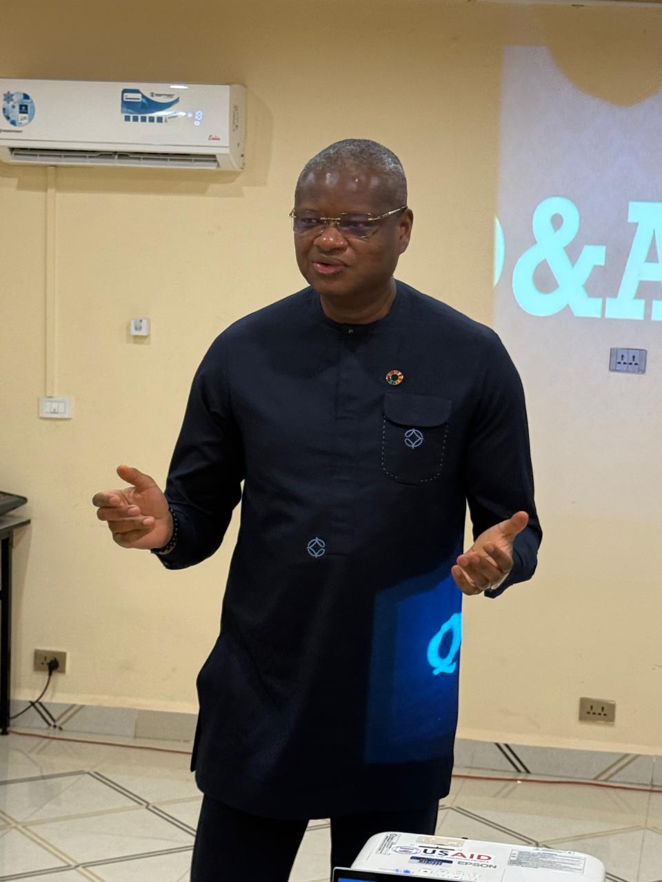 Presenter in a dark suit gesturing while speaking in a classroom with a projector screen.
