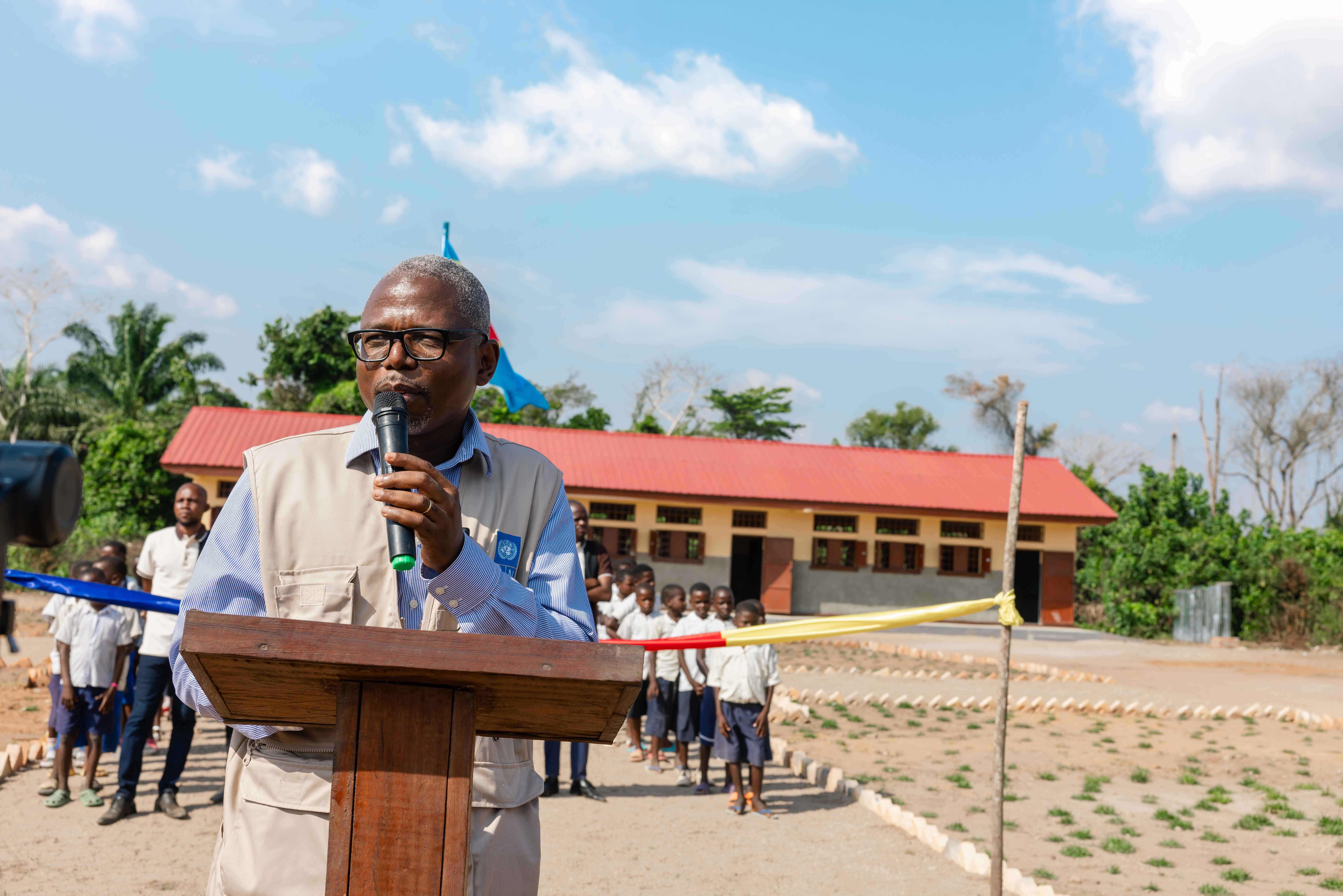 Photo: Man speaks at a wooden podium outdoors, with a crowd and building in the background.