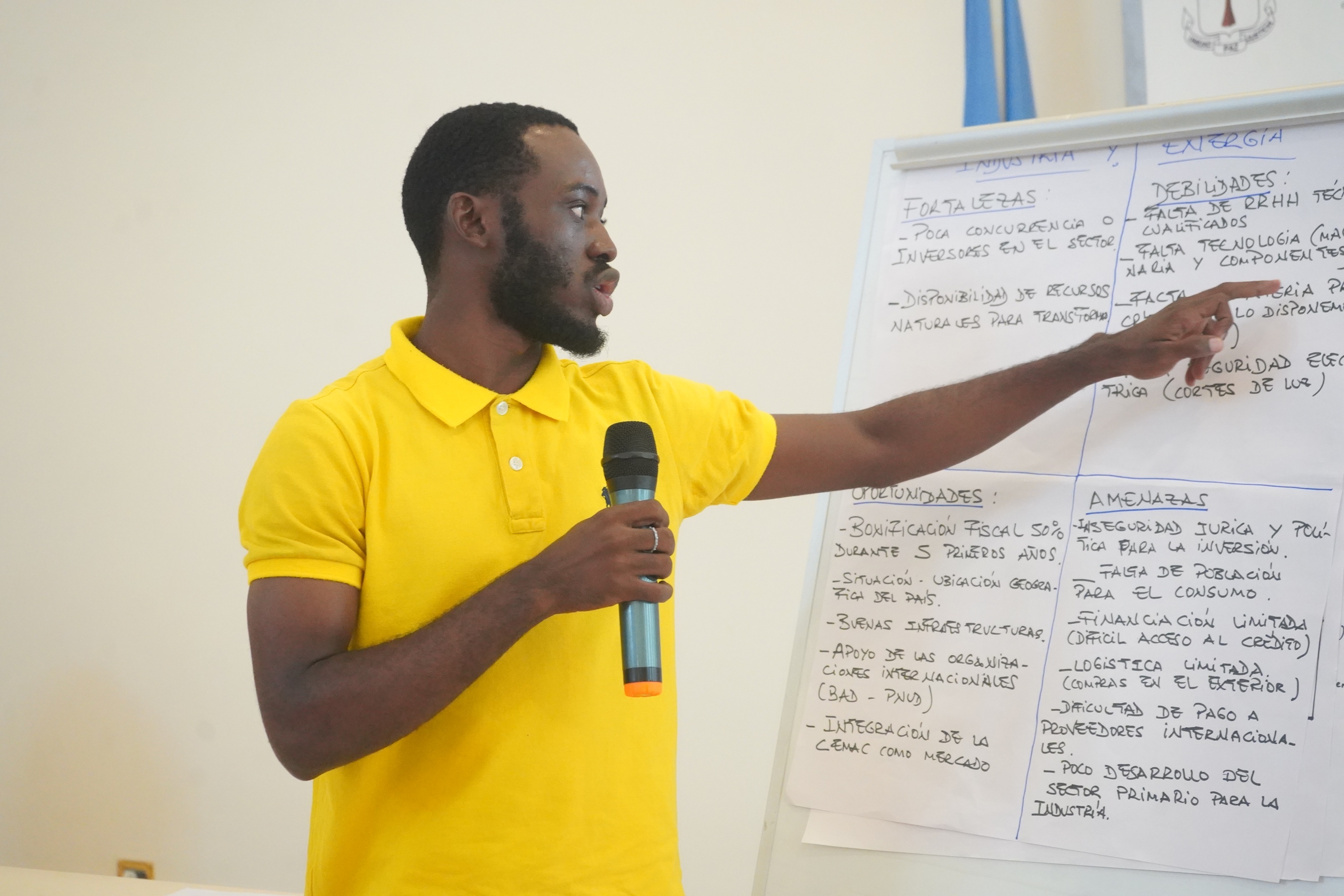 Man in yellow polo speaks, holding a microphone and pointing at a whiteboard.