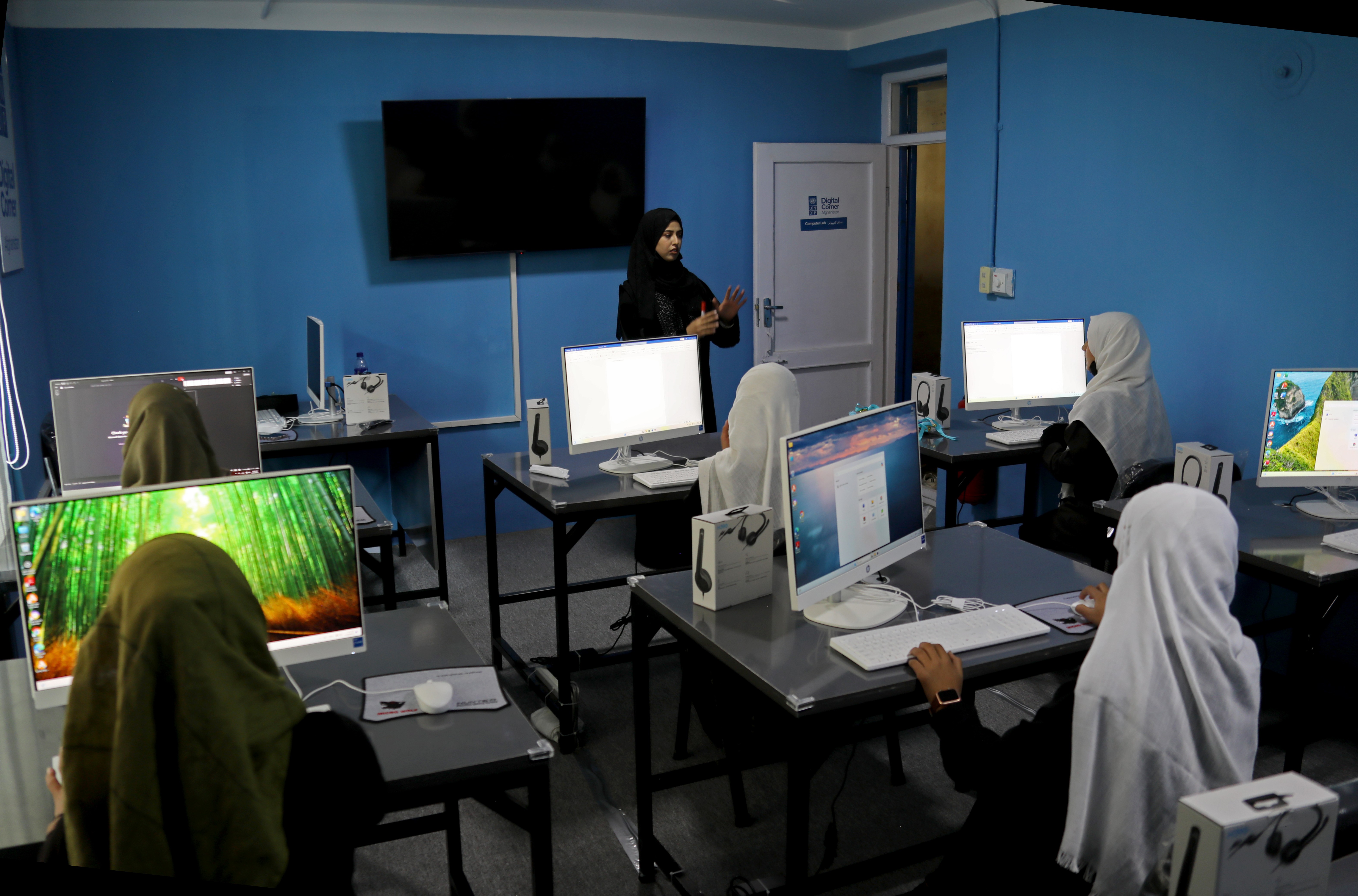 Photograph of blue-walled computer lab with students wearing hijabs at desks; instructor at front.