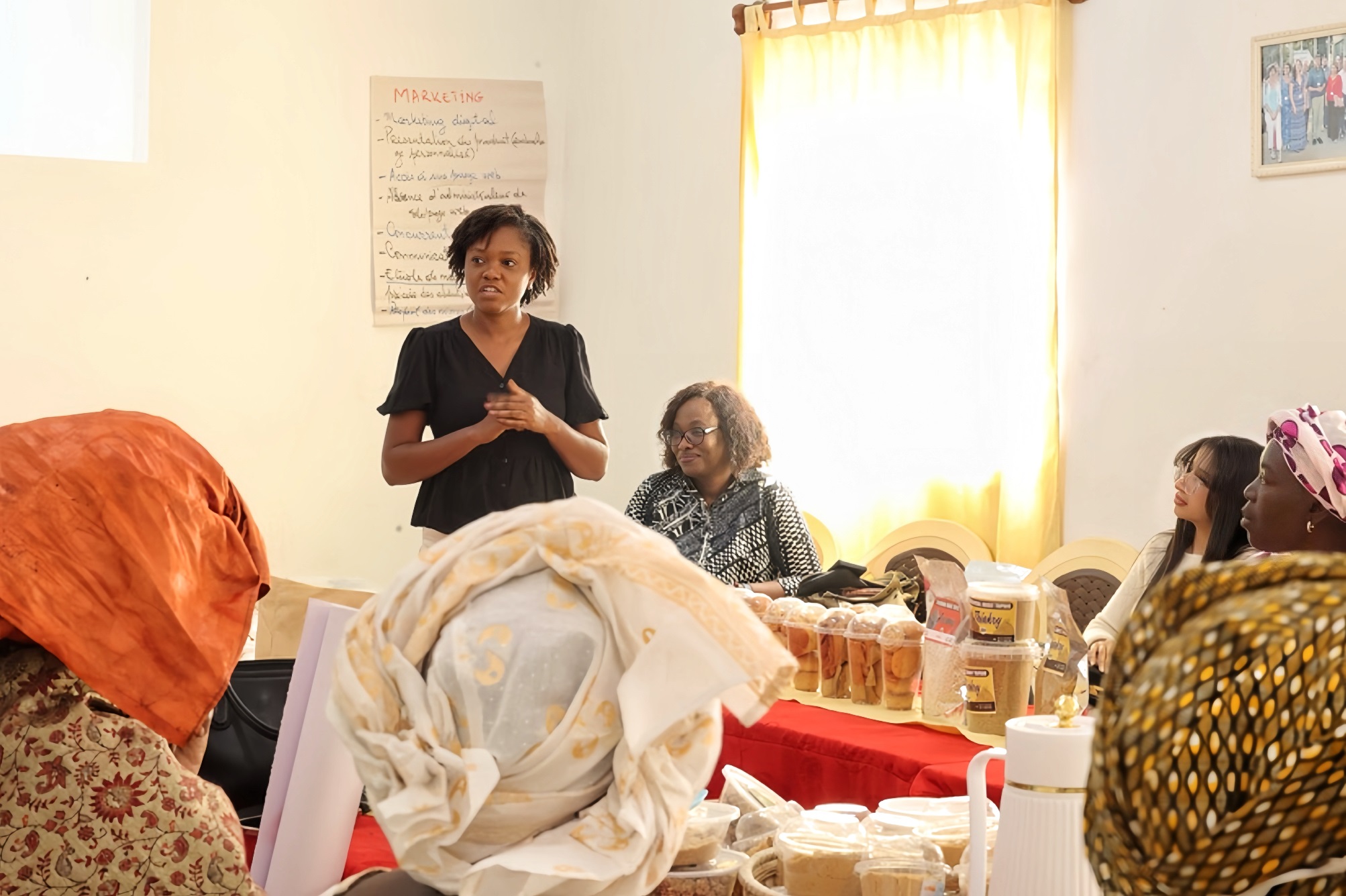 Group of women in a sunlit room; one woman stands and speaks to the seated group.