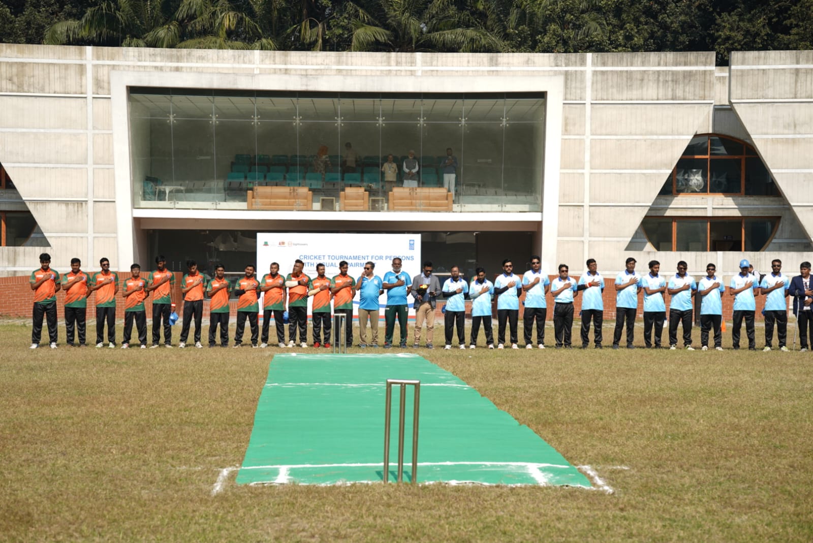 Group of students in orange and blue uniforms stand along a green field during an outdoor ceremony.