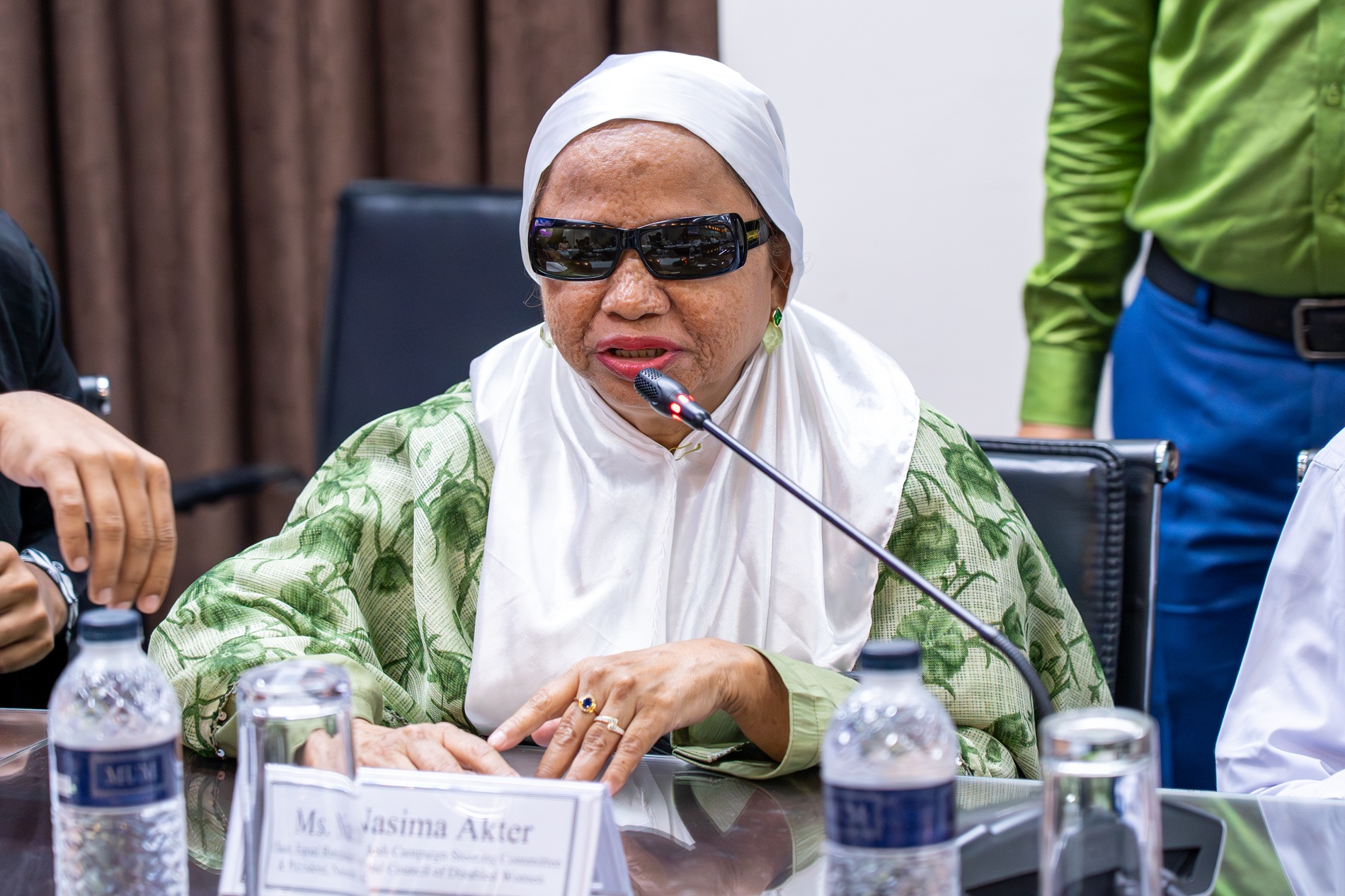 Woman in white hijab and green patterned blouse sits at a conference table with water bottles.