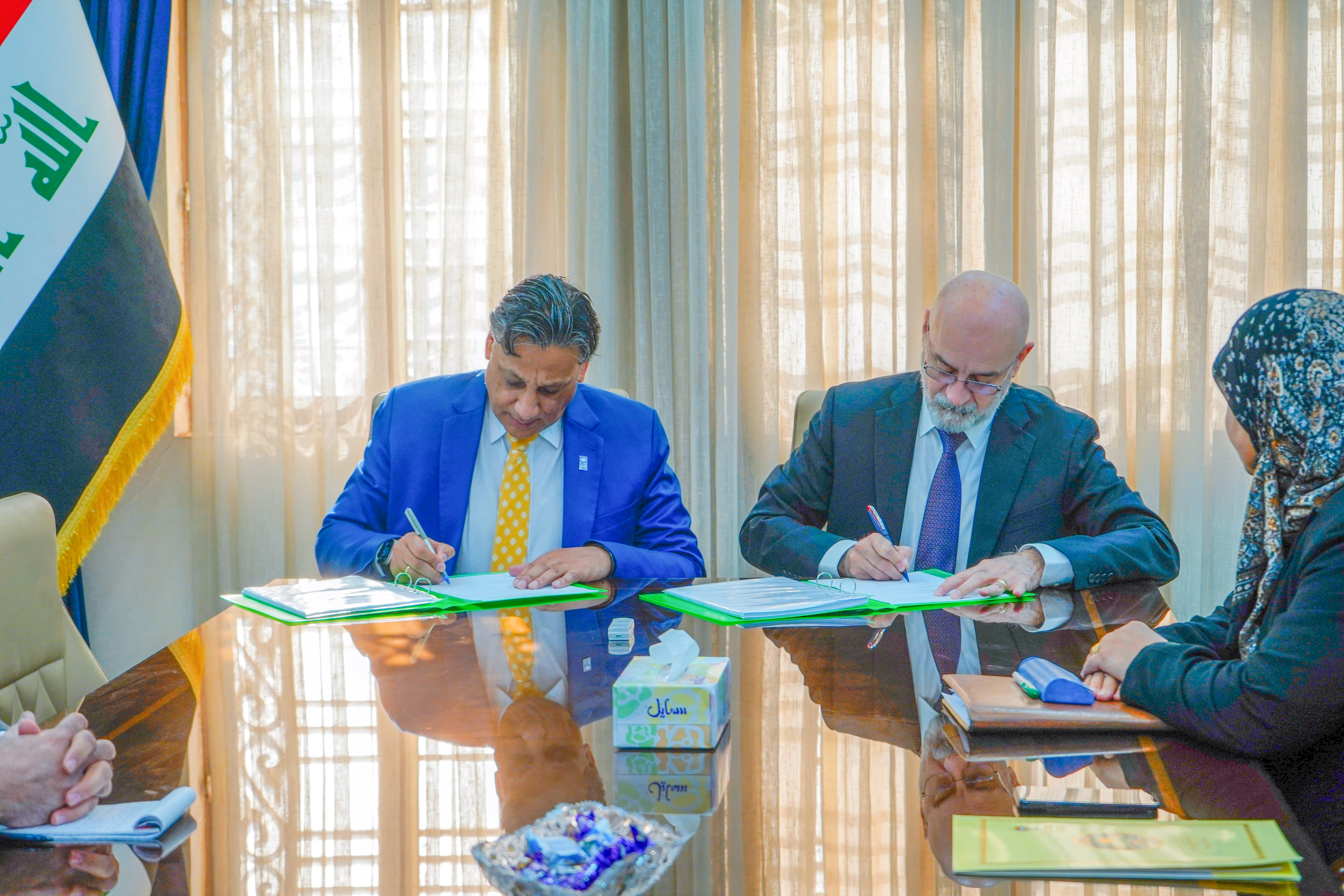 Two men and a woman sign documents at a glass table; flag in the background.