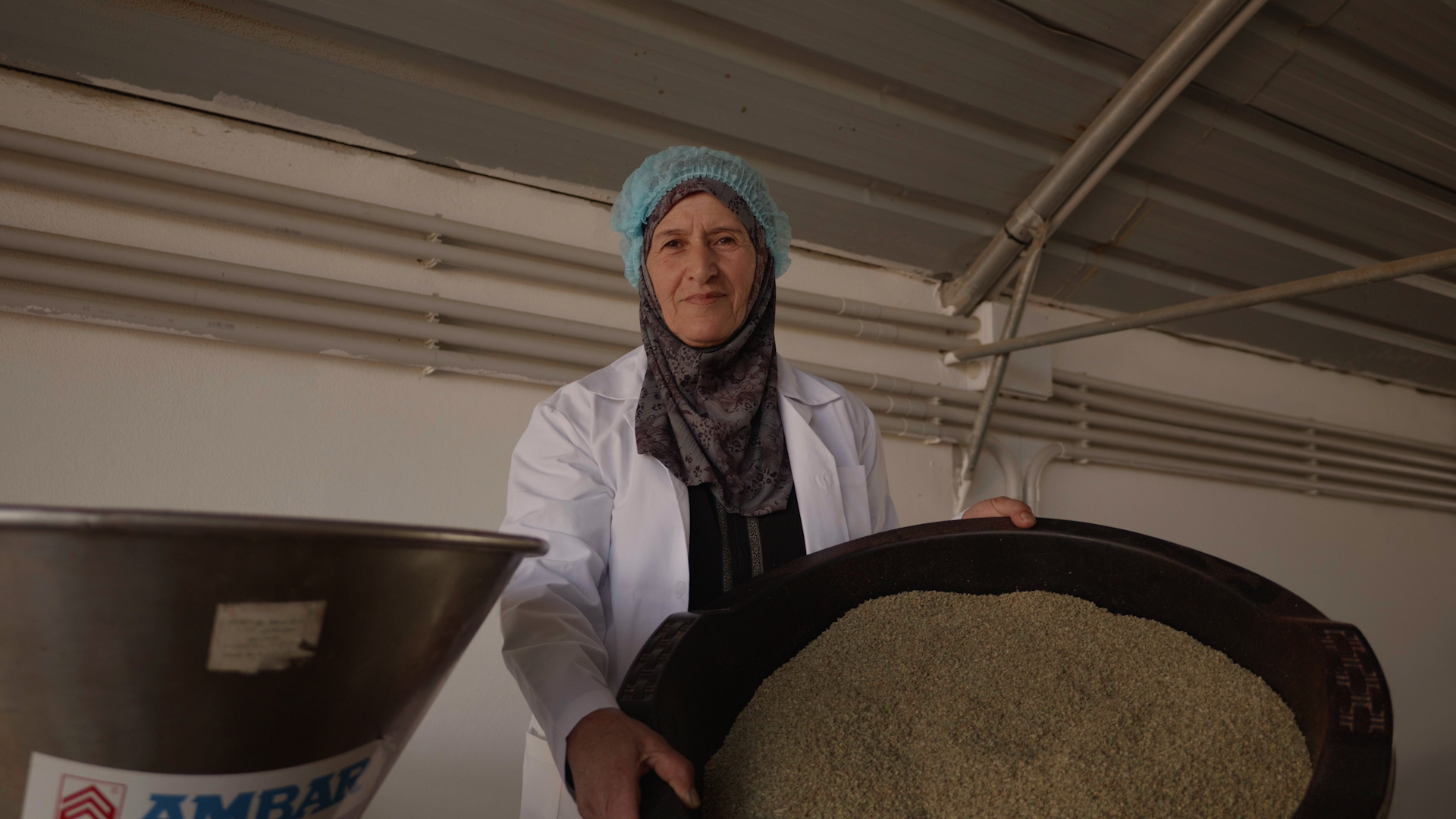 Worker in a white coat stands beside a large round tub filled with grain.